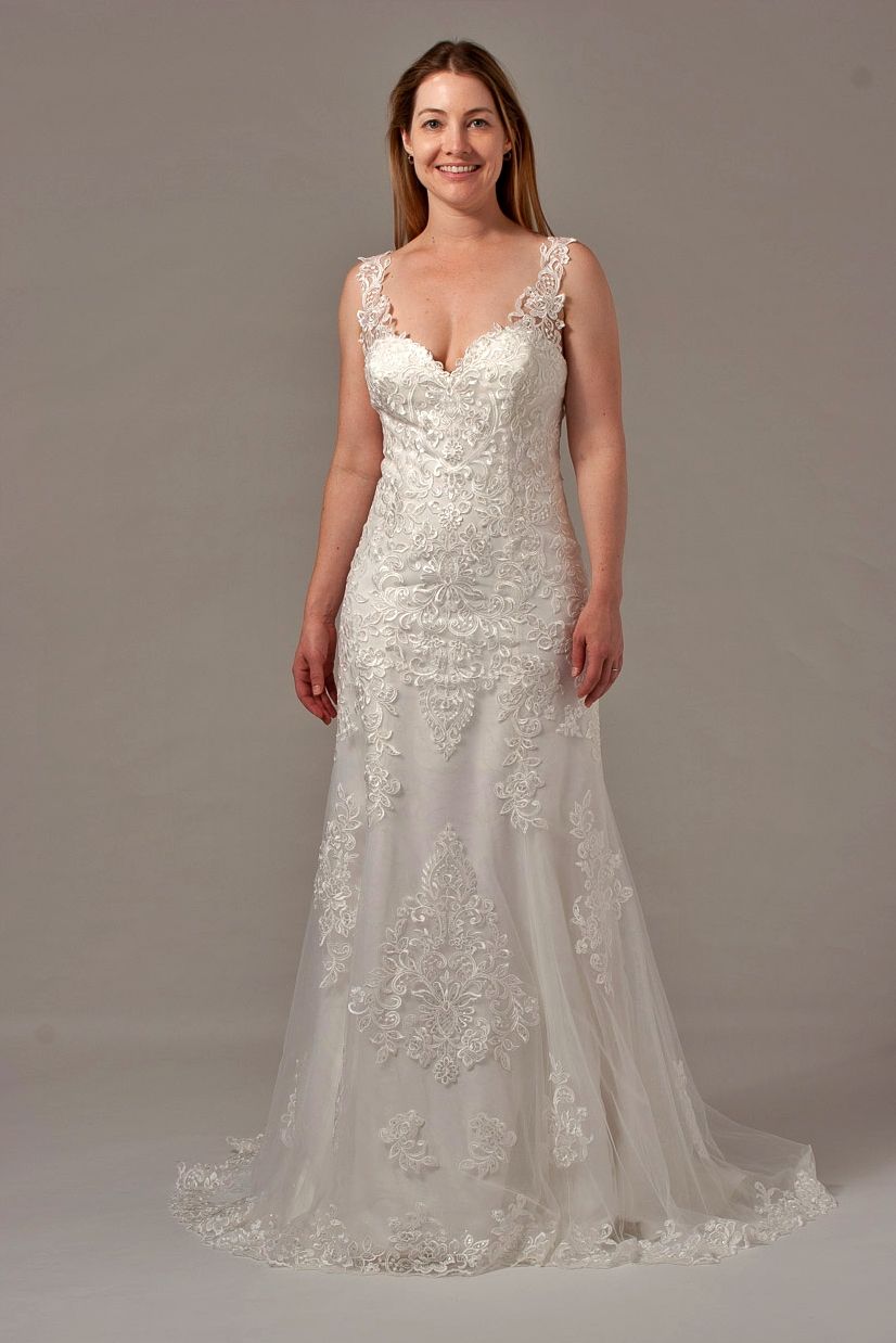 Woman in a white wedding dress with lace detail, smiling, posing in front of a gray backdrop.