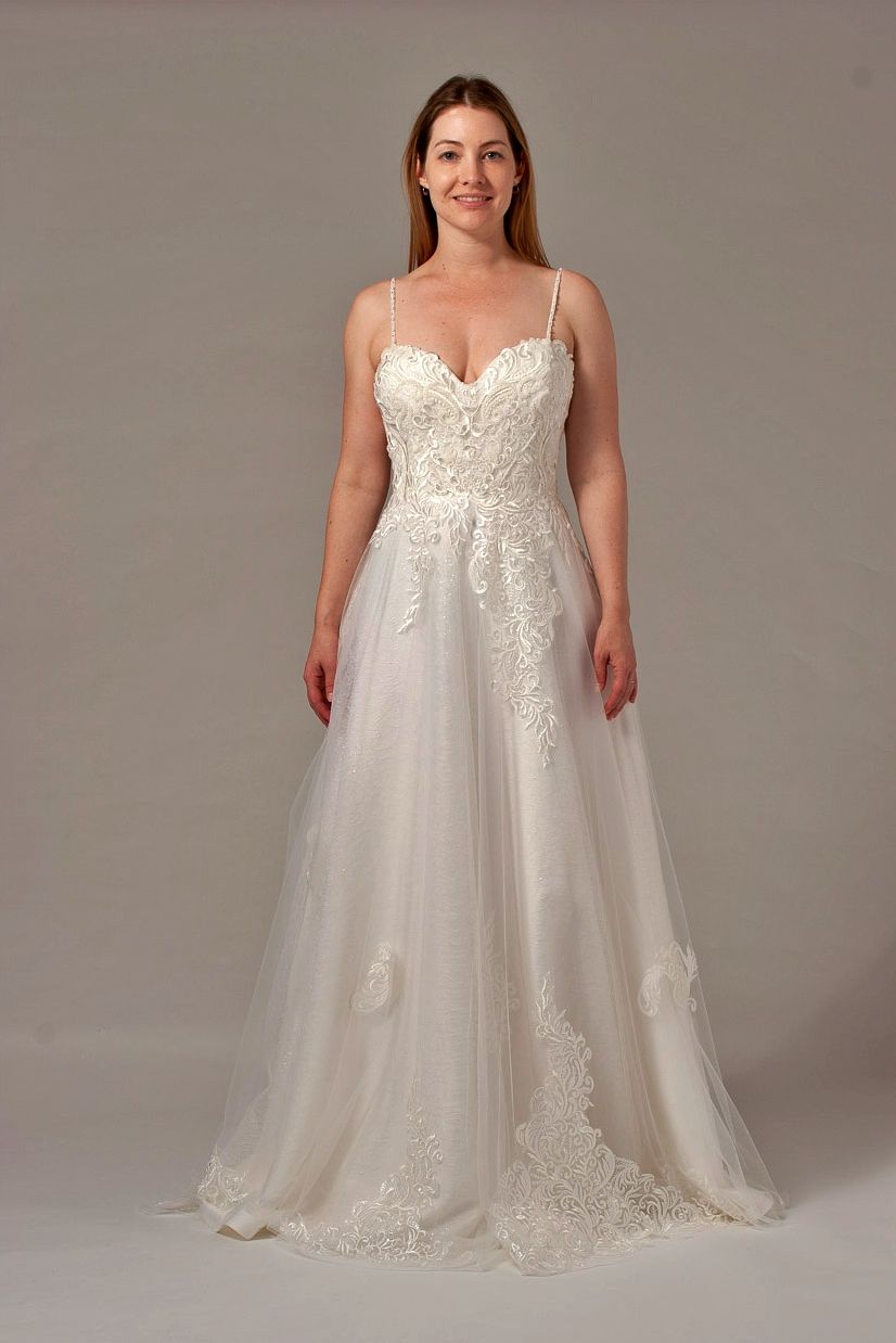 Woman wearing a white, embellished wedding dress, standing indoors, smiling.