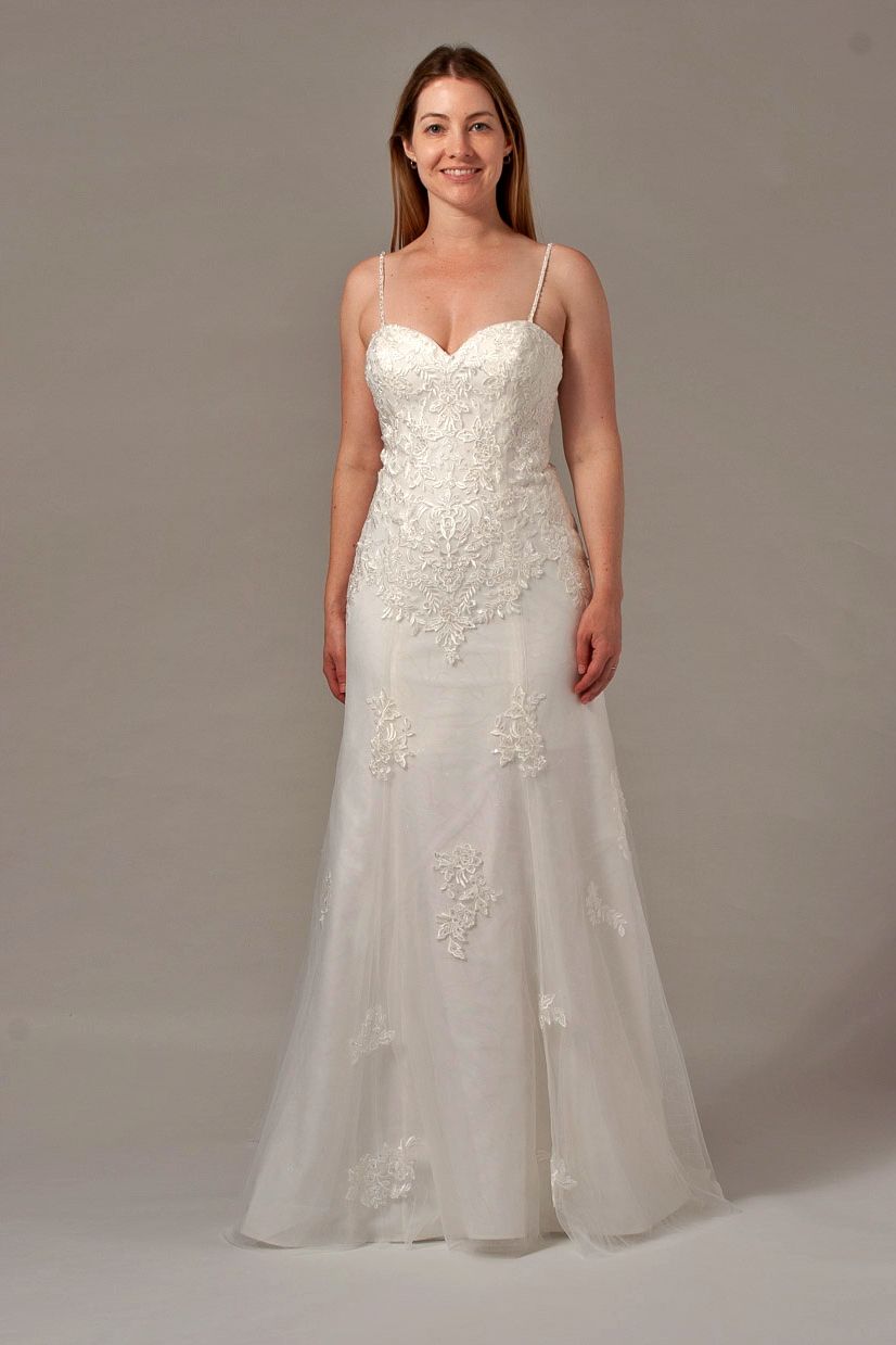 Woman in a white wedding dress with lace details, standing in a studio with a neutral background.