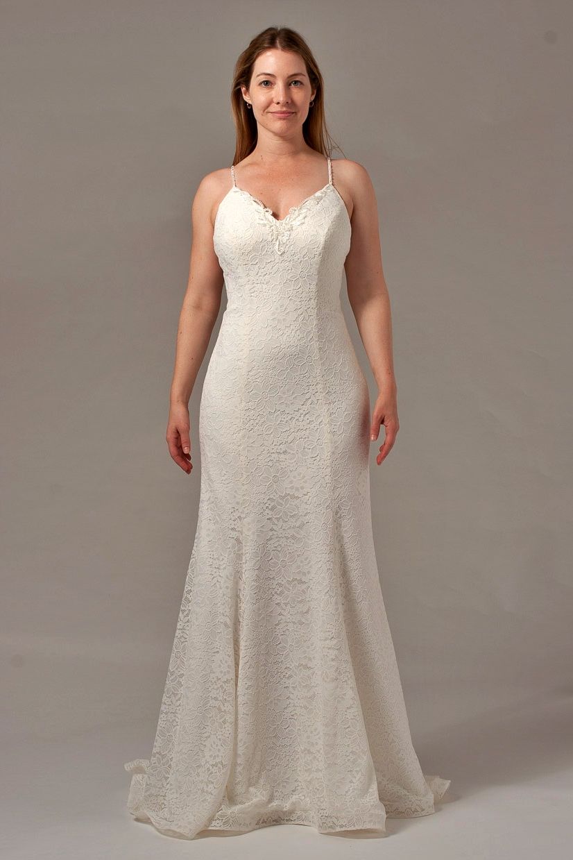 Woman in a cream-colored wedding dress with spaghetti straps, smiling, standing in a studio setting.