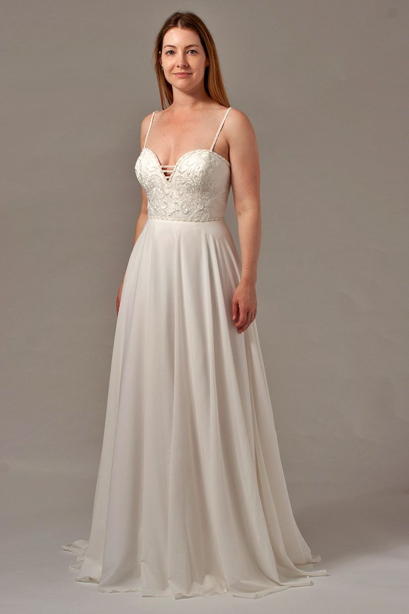 Woman in a white wedding dress with spaghetti straps, standing in front of a neutral backdrop.