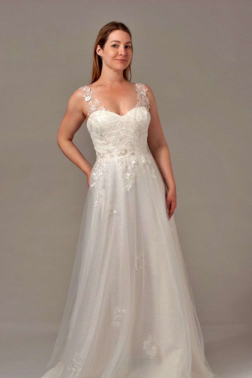 Woman in a white wedding dress with floral bodice and a-line skirt, standing with a hand in her pocket.