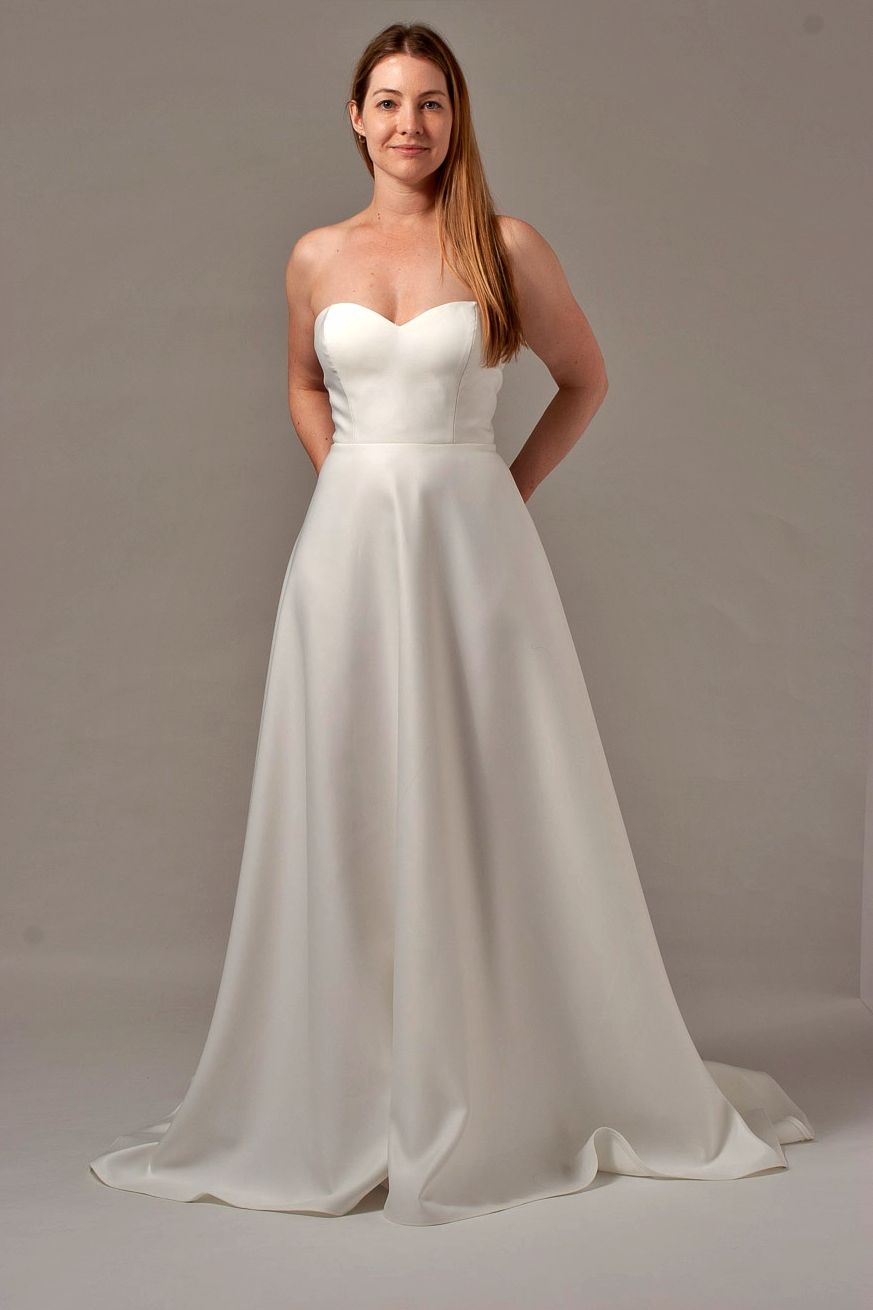 Woman in a white strapless wedding dress, standing with hands behind her back, against a grey background.