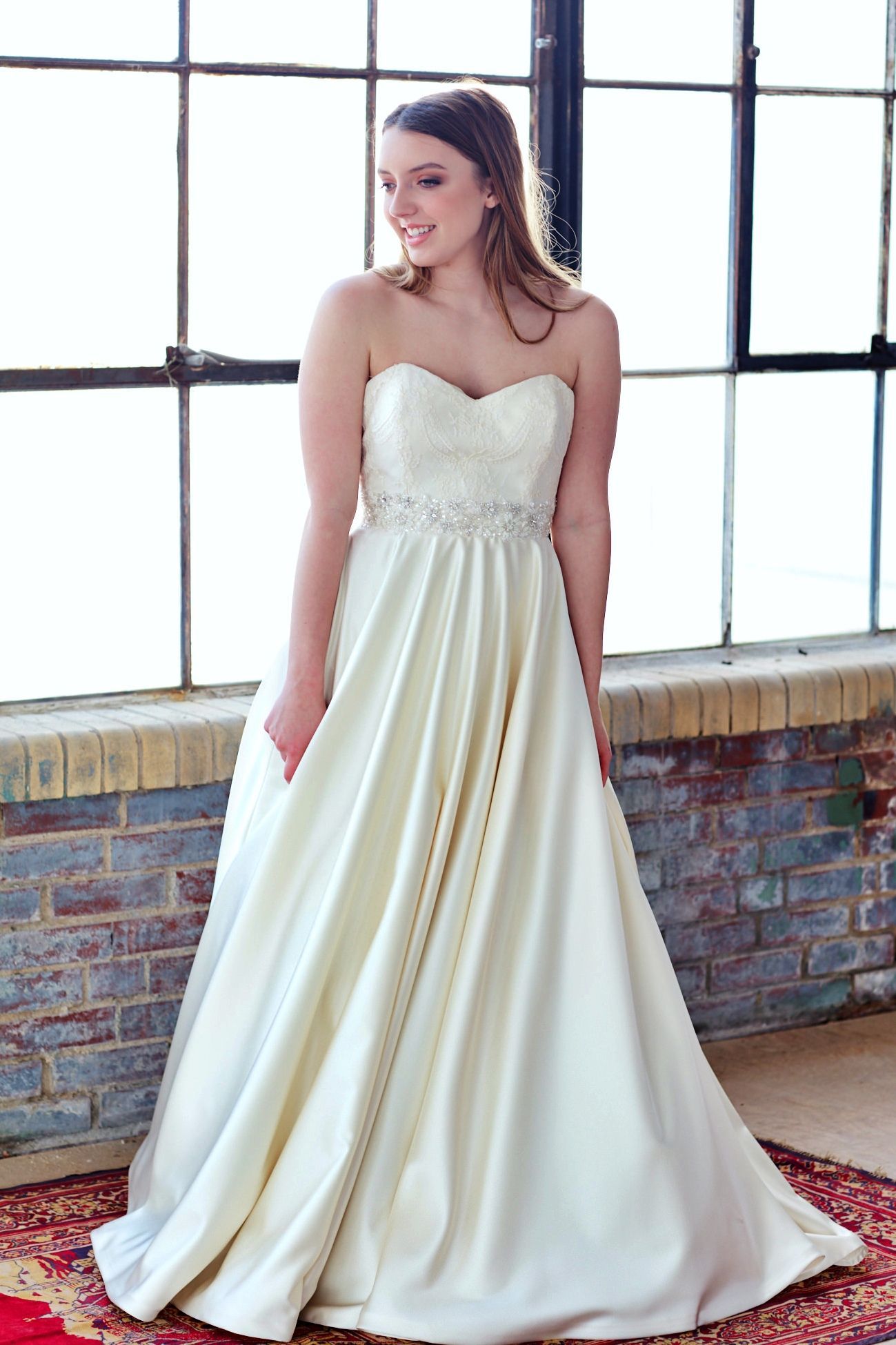 Woman in a strapless ivory wedding gown stands by a window, smiling.