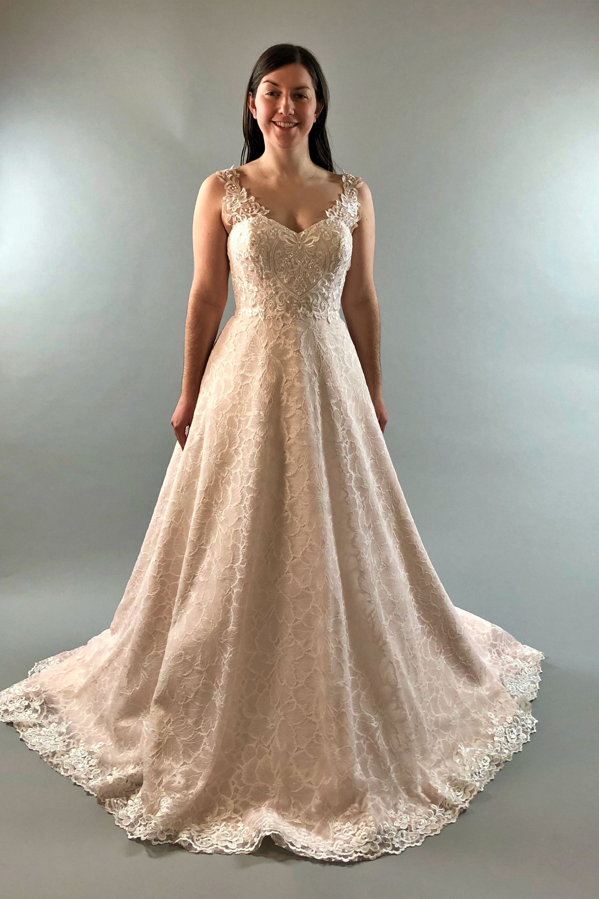 Woman wearing a blush-colored wedding dress with beaded bodice, A-line skirt, and lace trim, smiling.