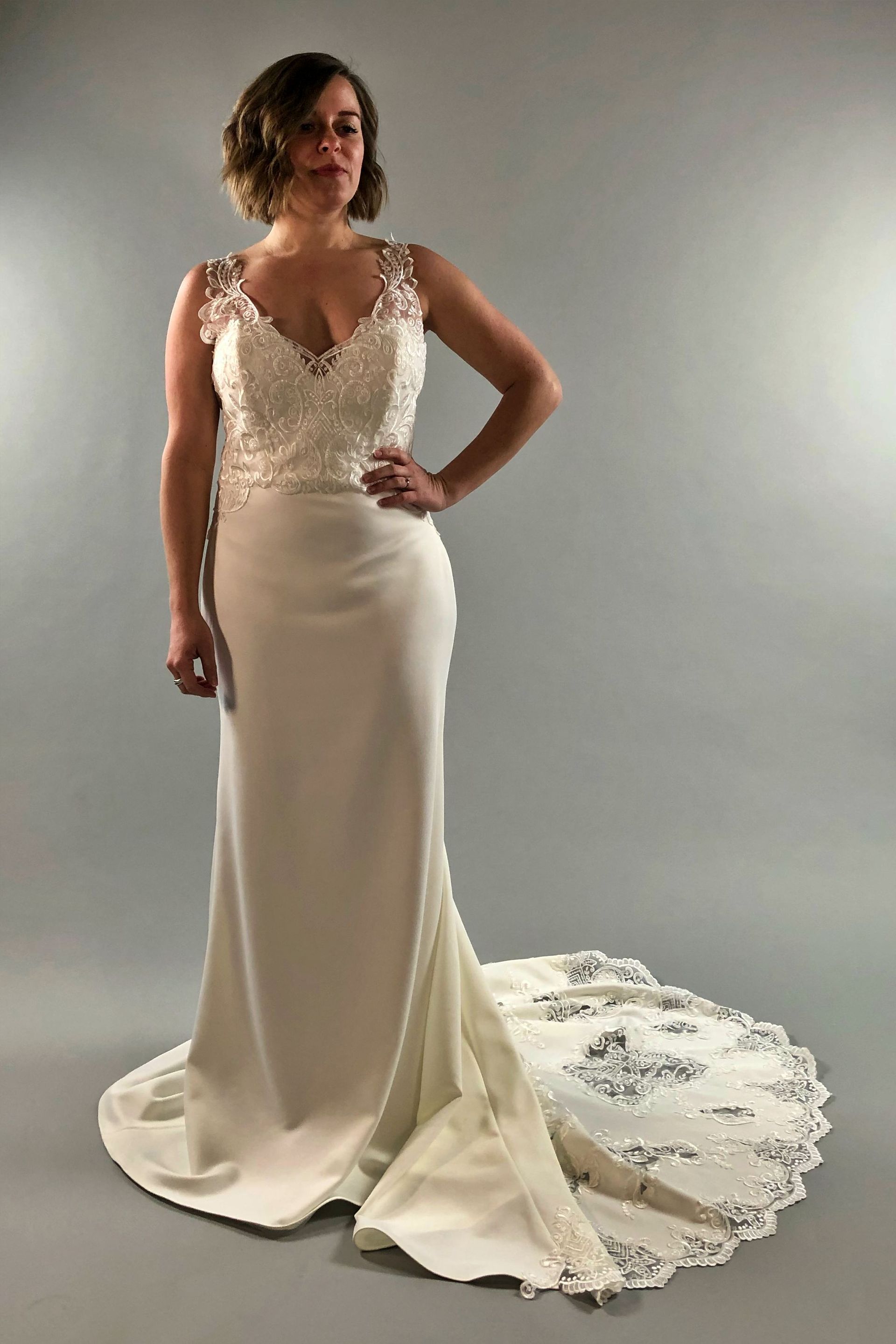 Woman in a white wedding dress with a decorated bodice and train, standing with hand on hip.