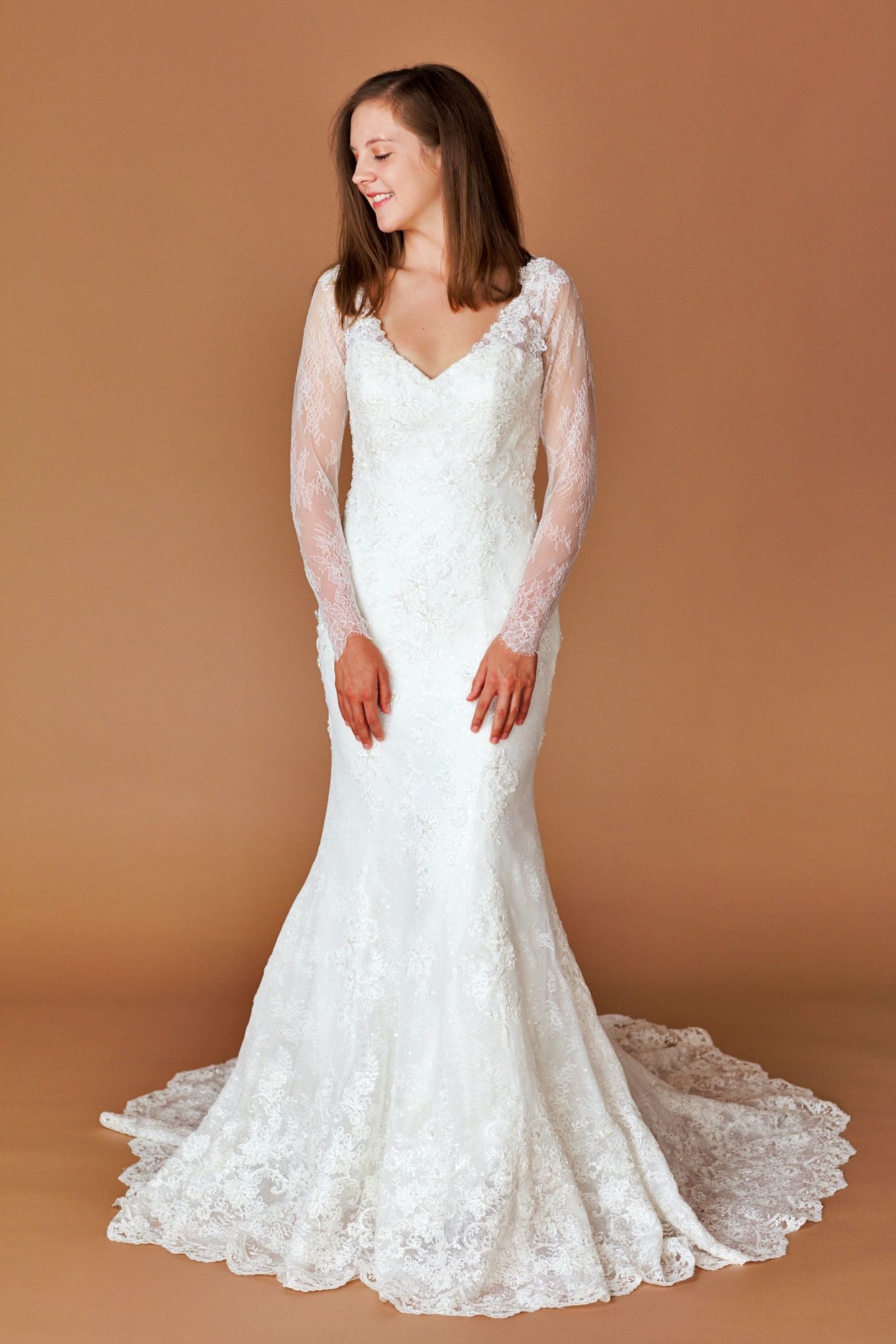 Woman in a white lace wedding dress with long sleeves, smiling, posing against a light brown backdrop.
