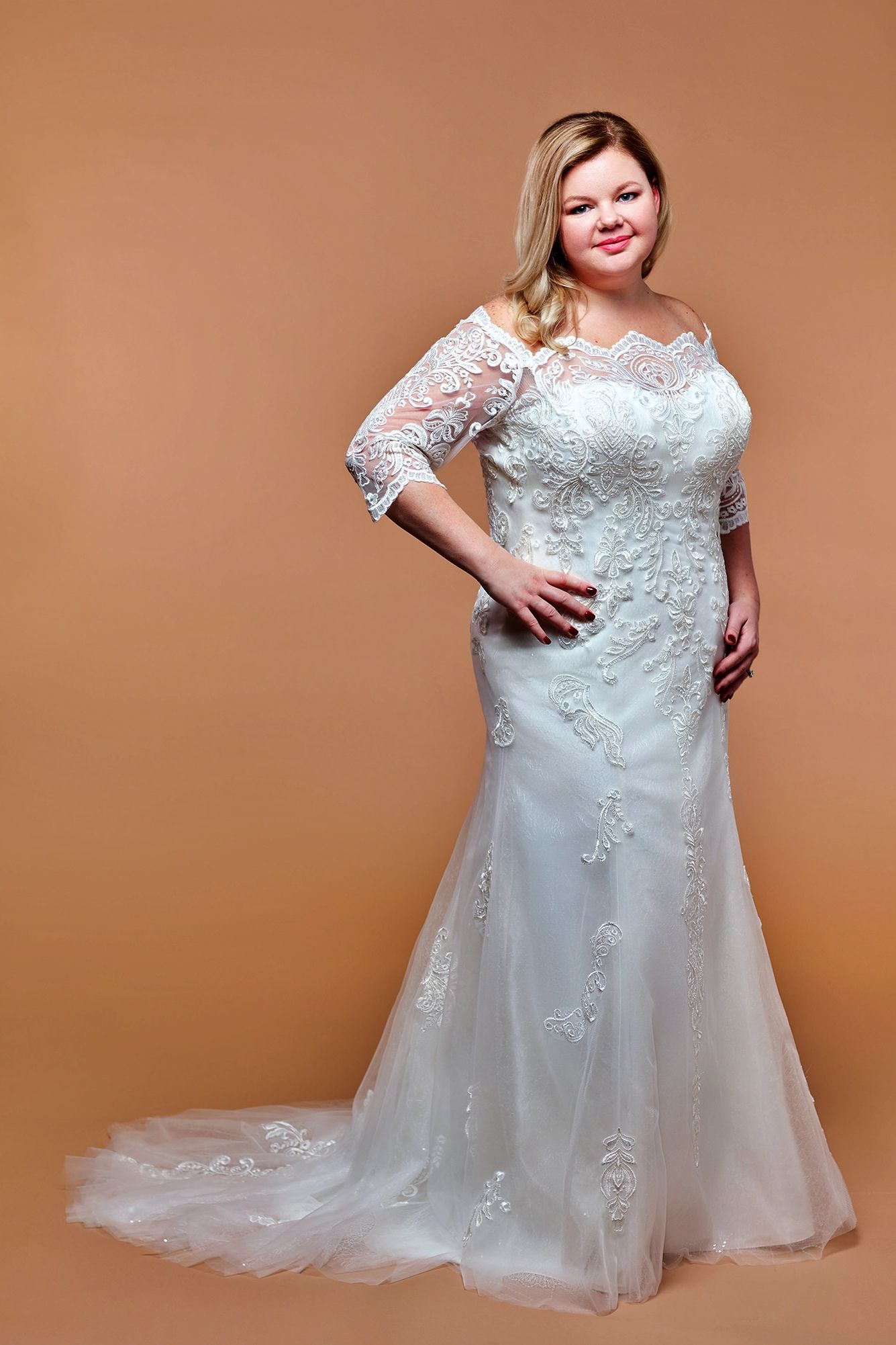 Woman in a white lace wedding dress with a train, standing against a beige backdrop, smiling.