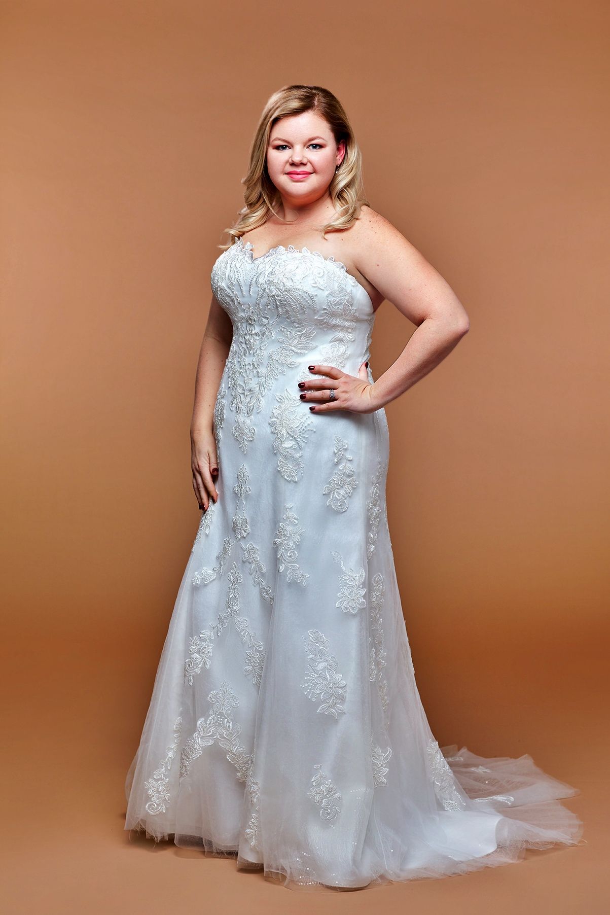 Woman in a strapless white wedding dress with lace details and a small train, posing.