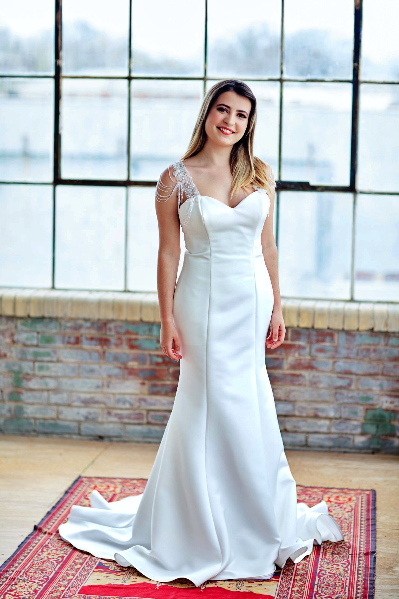 Woman in white wedding dress smiles in front of large window.