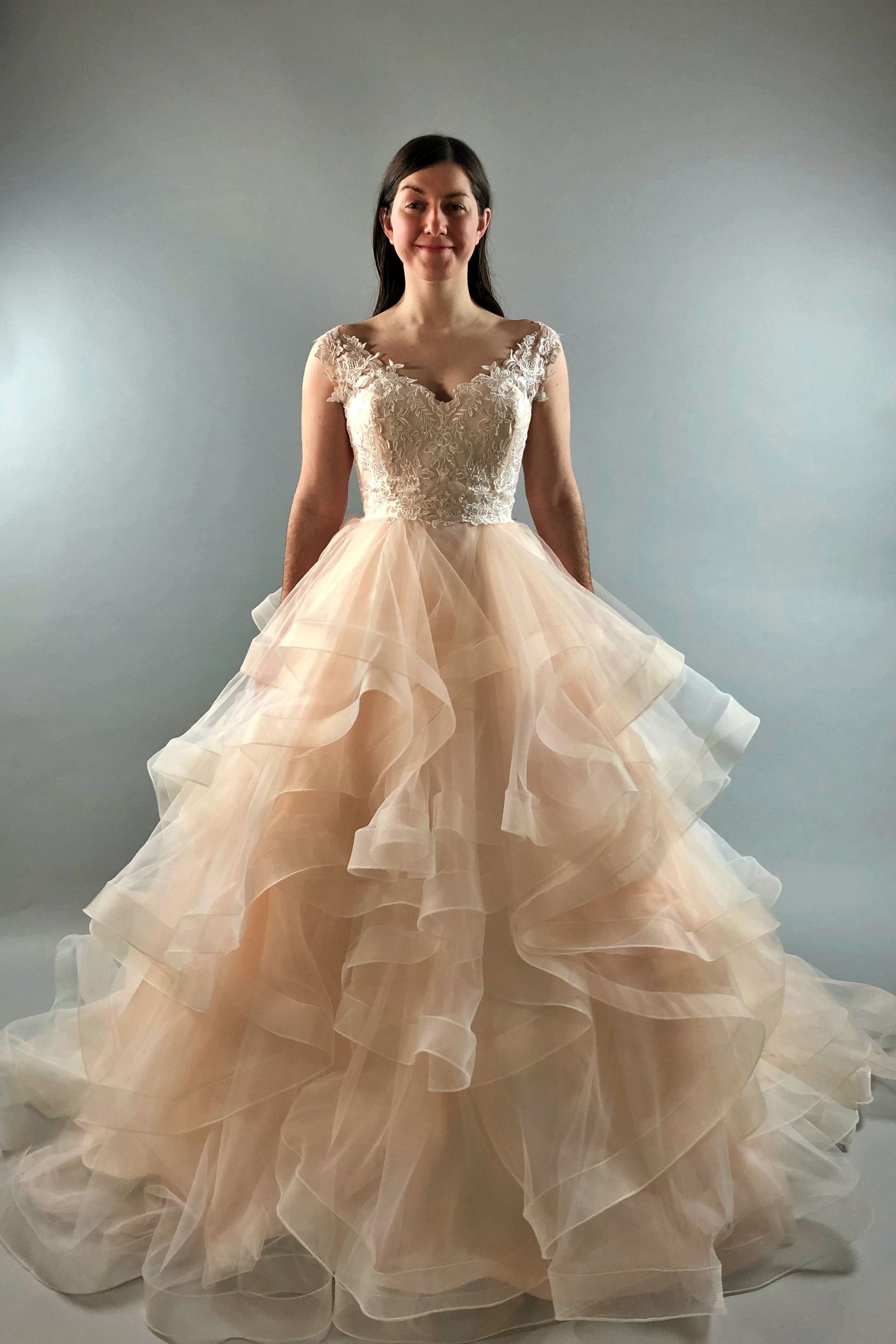 Woman in a peach-colored, tiered ballgown with jeweled bodice, standing and smiling in a studio.