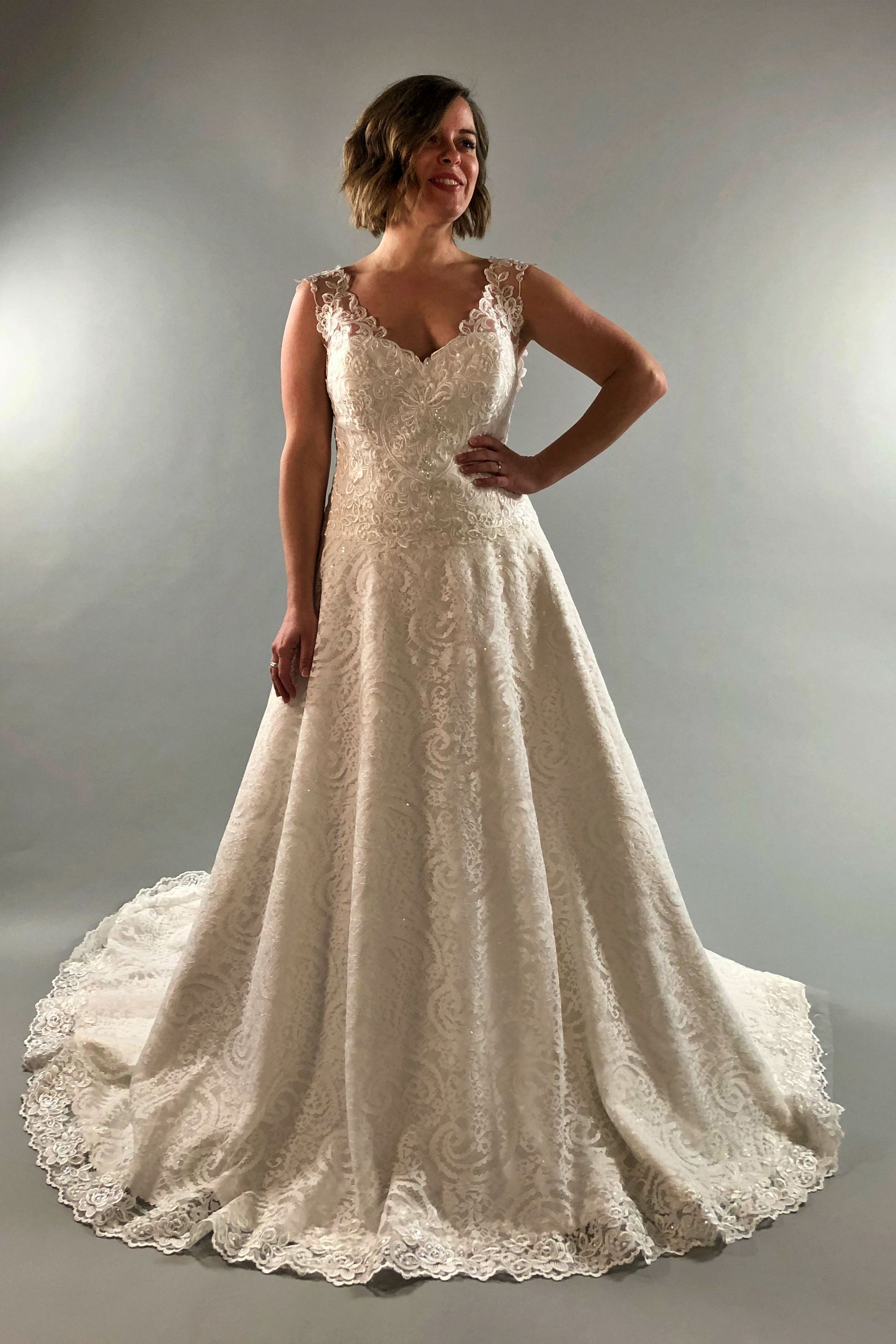 Woman in a white lace wedding gown with a flowing train. She is smiling and has one hand on her hip.
