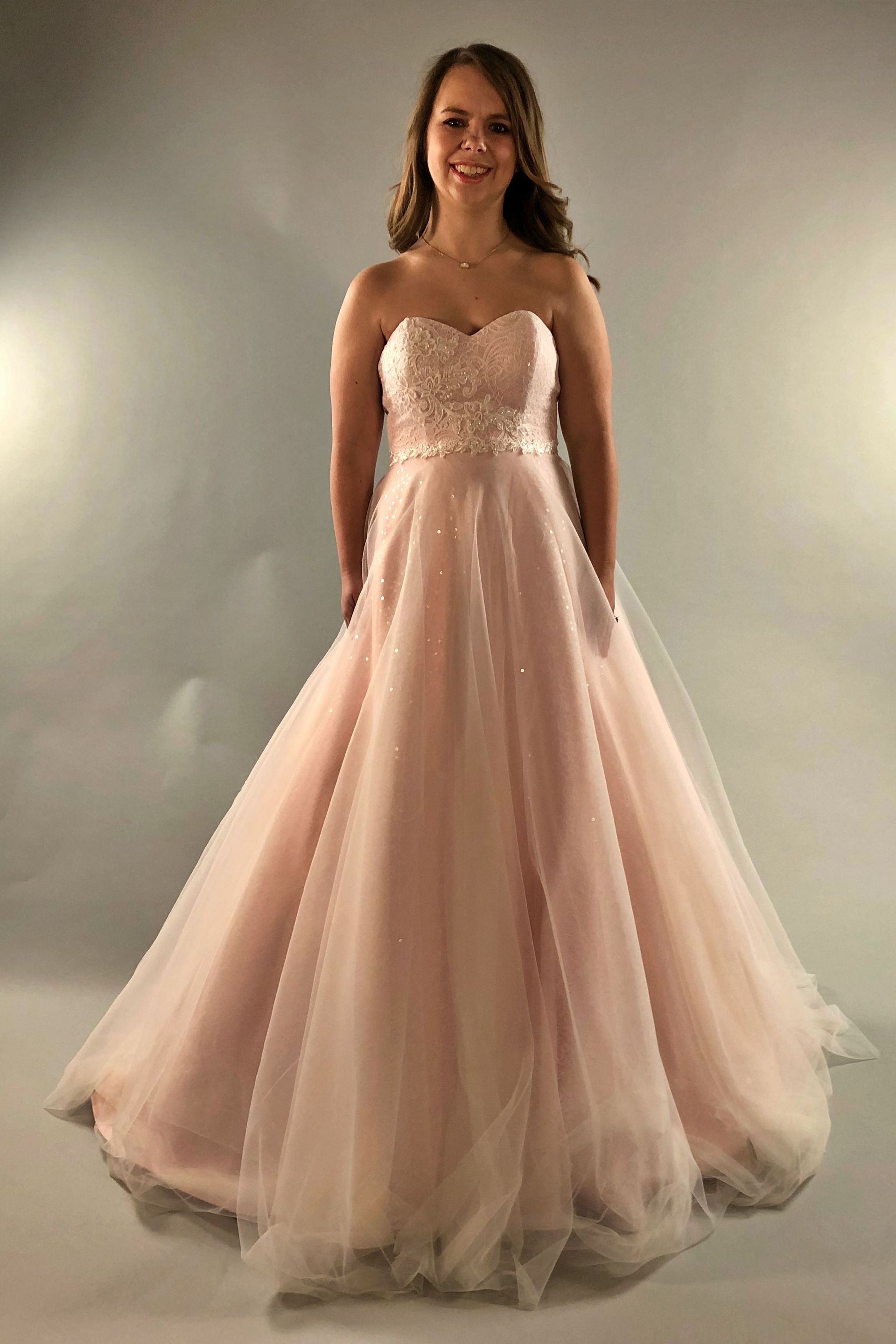 Woman in a blush pink strapless gown smiles, hands in pockets.