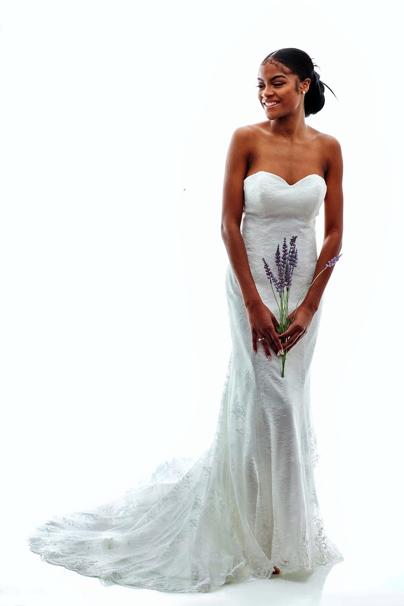 Woman in white strapless wedding gown, holding lavender, smiling; train, white background.