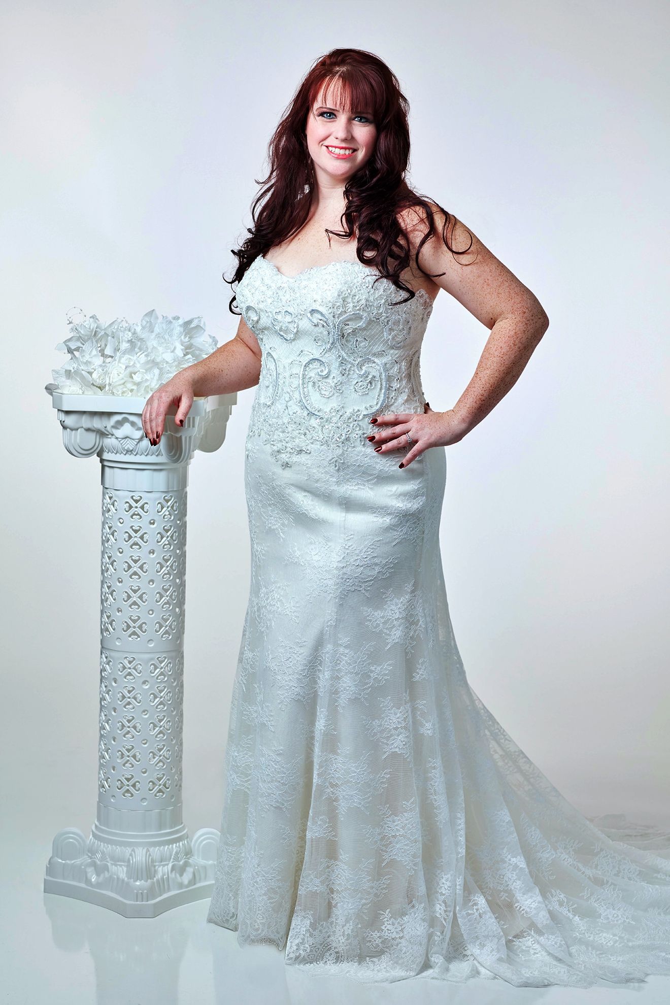 Woman in white wedding dress, smiling, leaning on ornate white column.