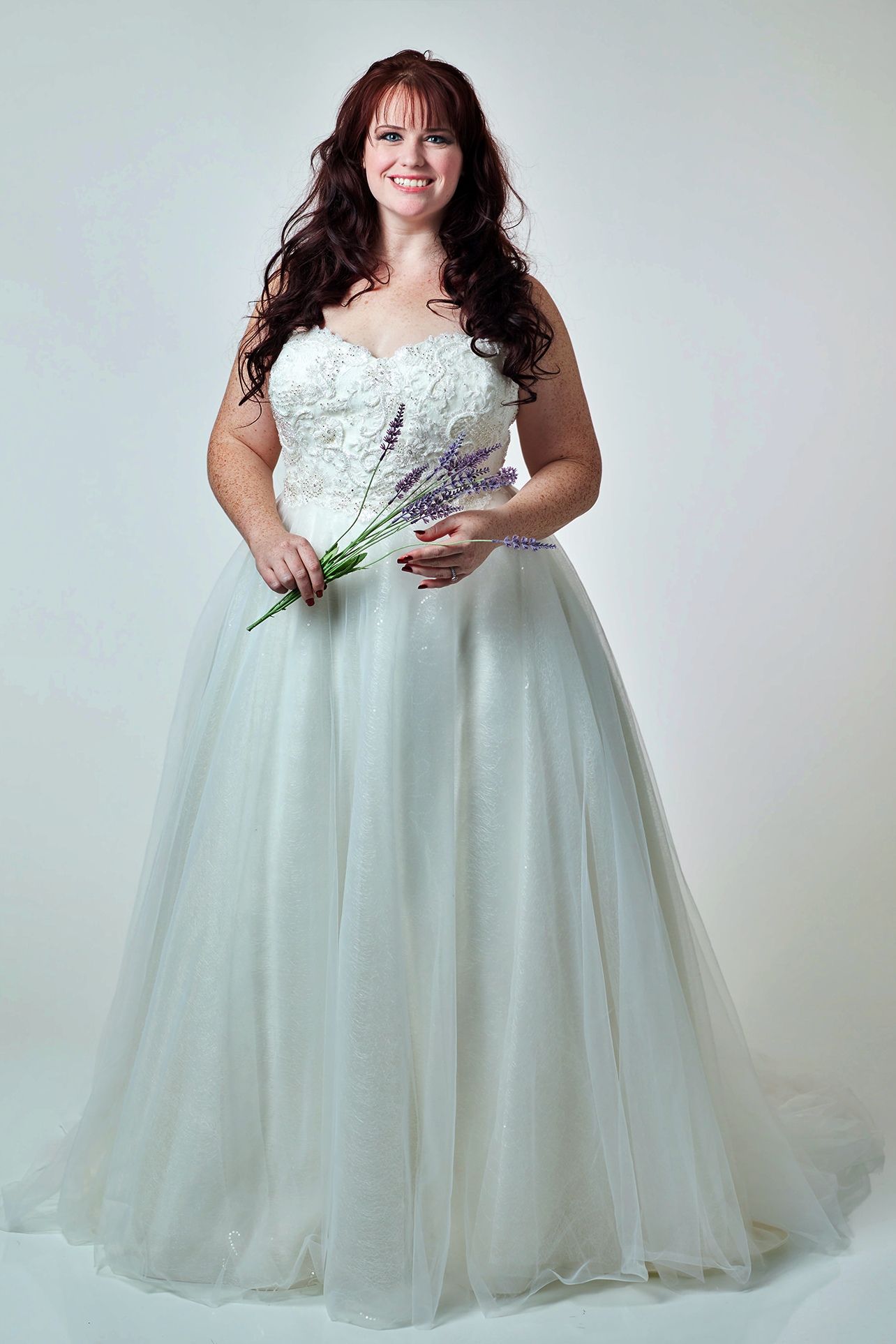 Woman in a white wedding gown, holding flowers, smiles, against a white background.