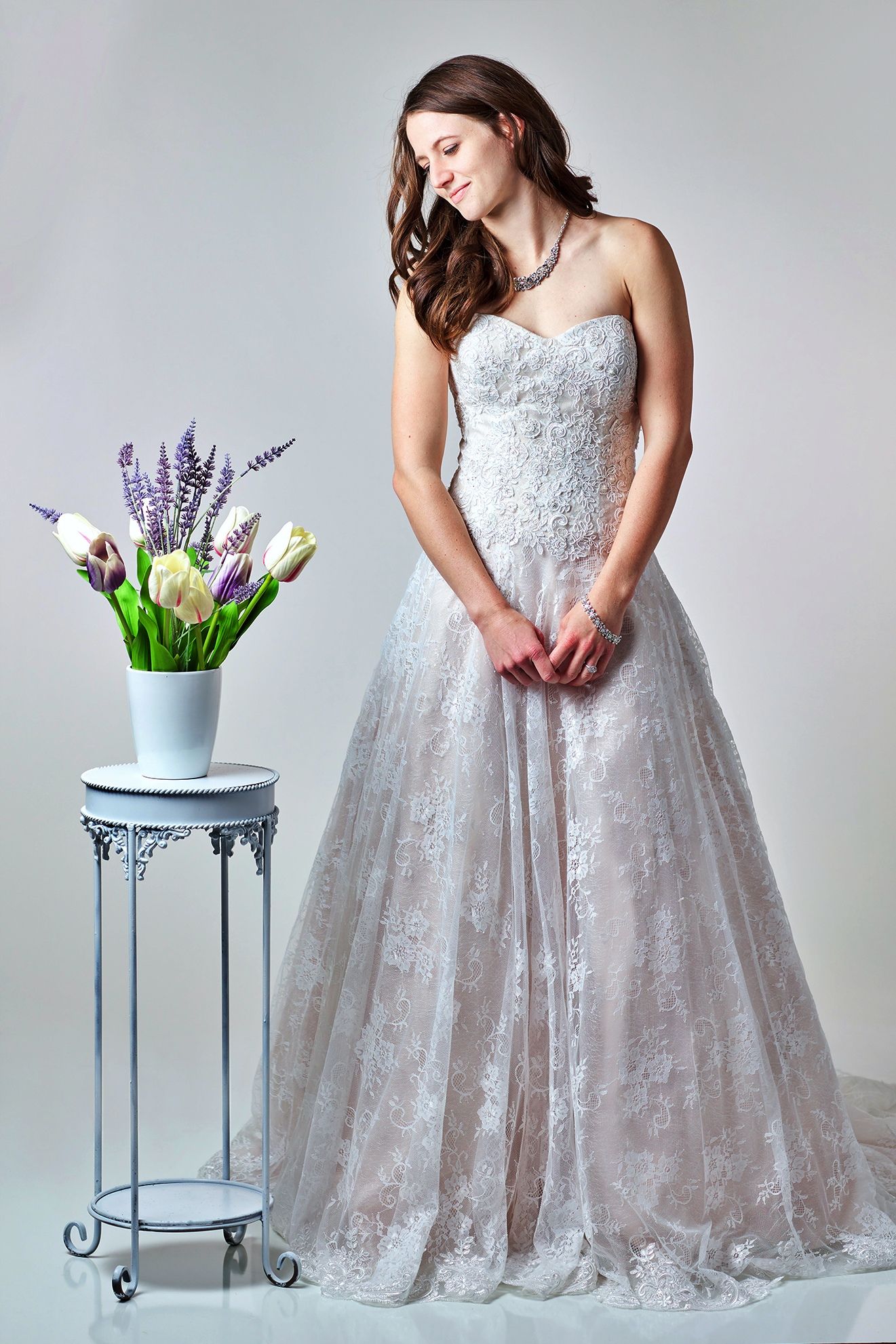 Woman in a white strapless wedding dress, smiling, next to a vase of flowers on a small table.