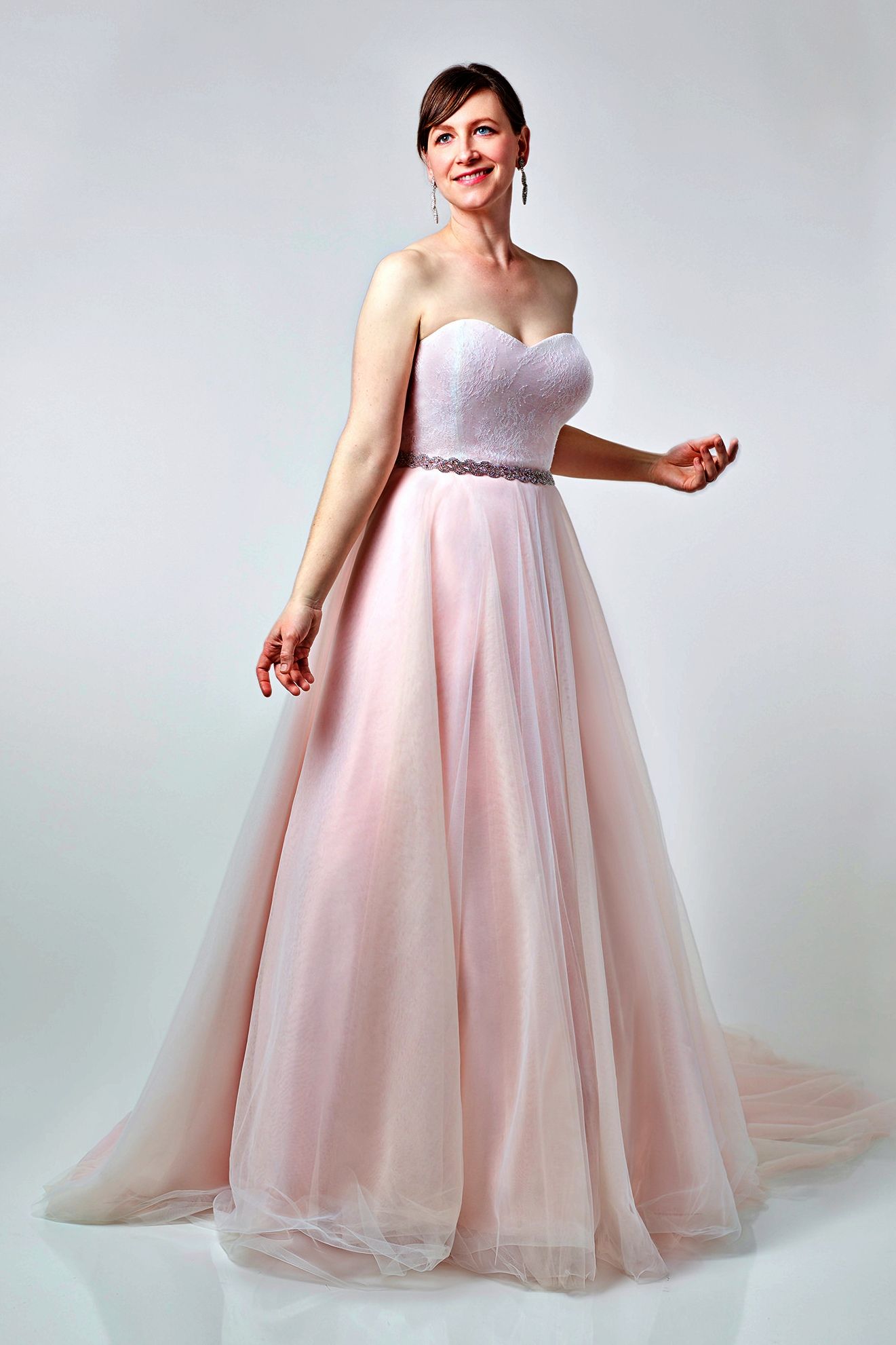 Woman in a strapless pink wedding gown, smiling, arms out. Light background.