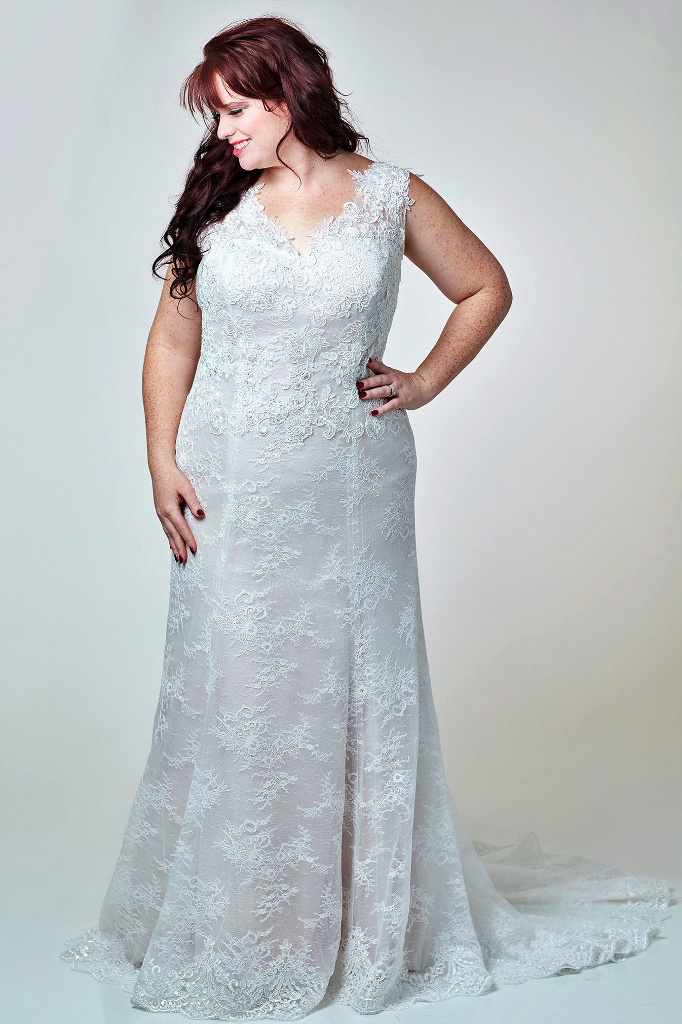 Woman in a white lace wedding dress, smiling. She has one hand on her hip and is in a studio.