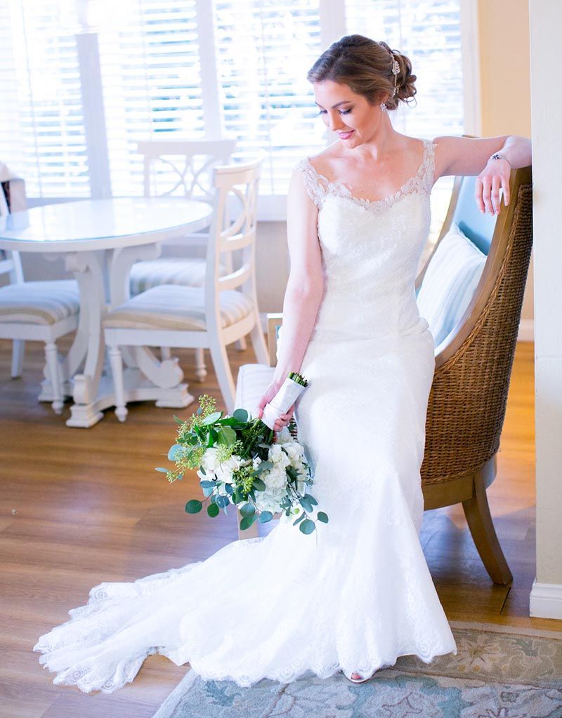 Bride in white gown, holding bouquet, leaning against wicker chair in a bright room.
