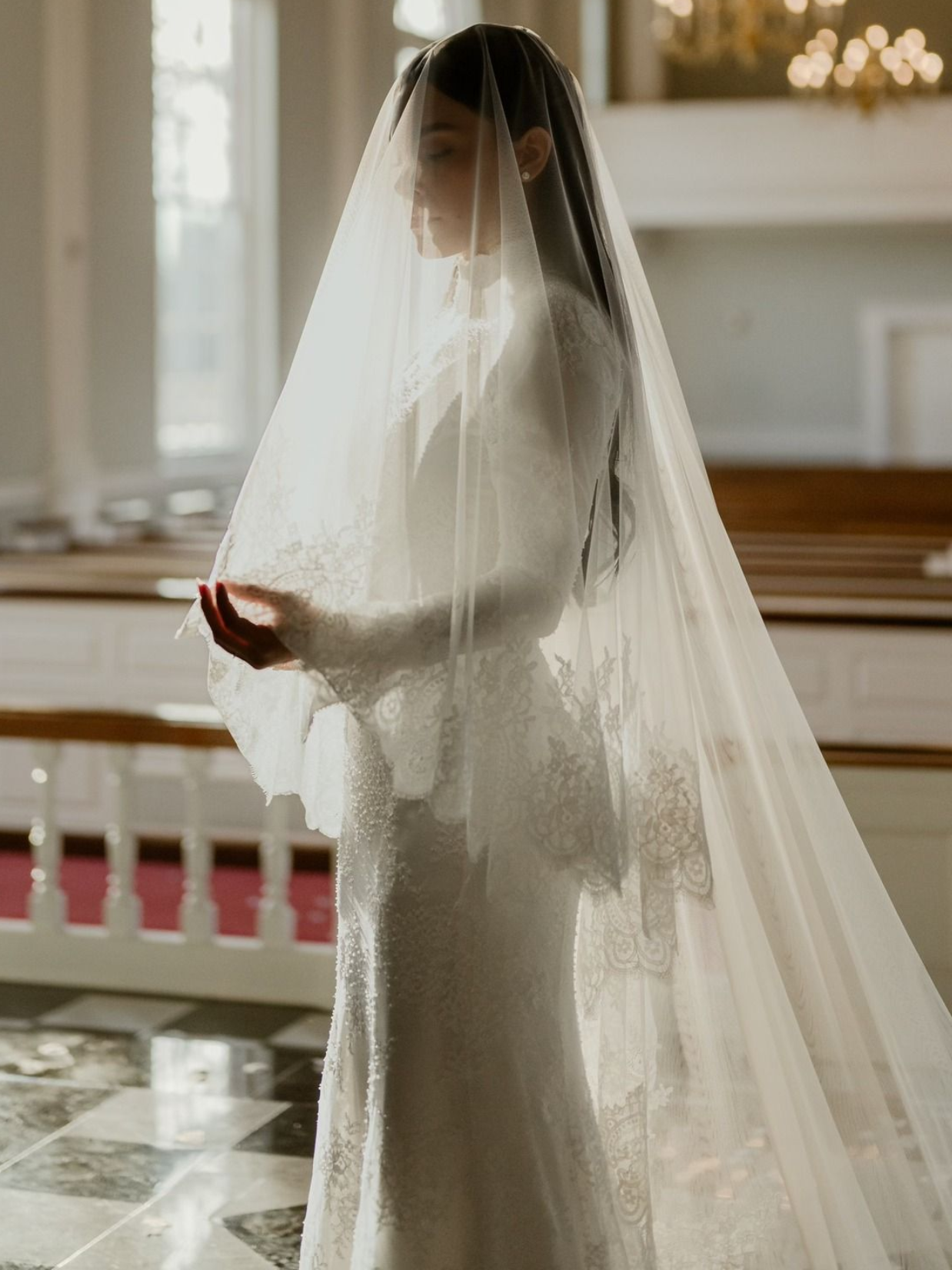 Two women in white wedding dresses, smiling, holding hands, looking at each other against a white background.