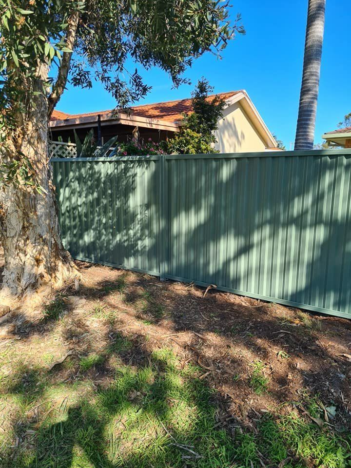 A Green Fence Is Surrounded by Grass and Trees in Front of A House — D & B Fencing in Taree, NSW