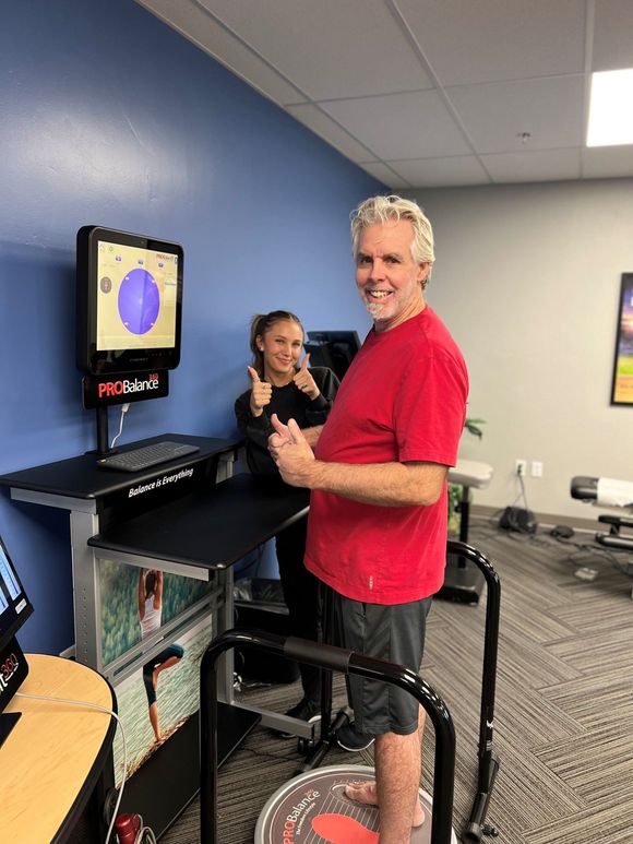 Person in a red shirt standing at an exercise station in a gym, smiling at the camera.