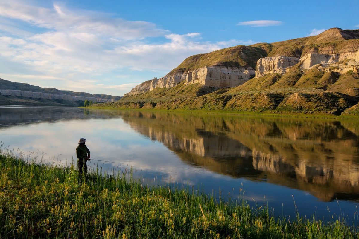 A man is fishing in a lake with mountains in the background.