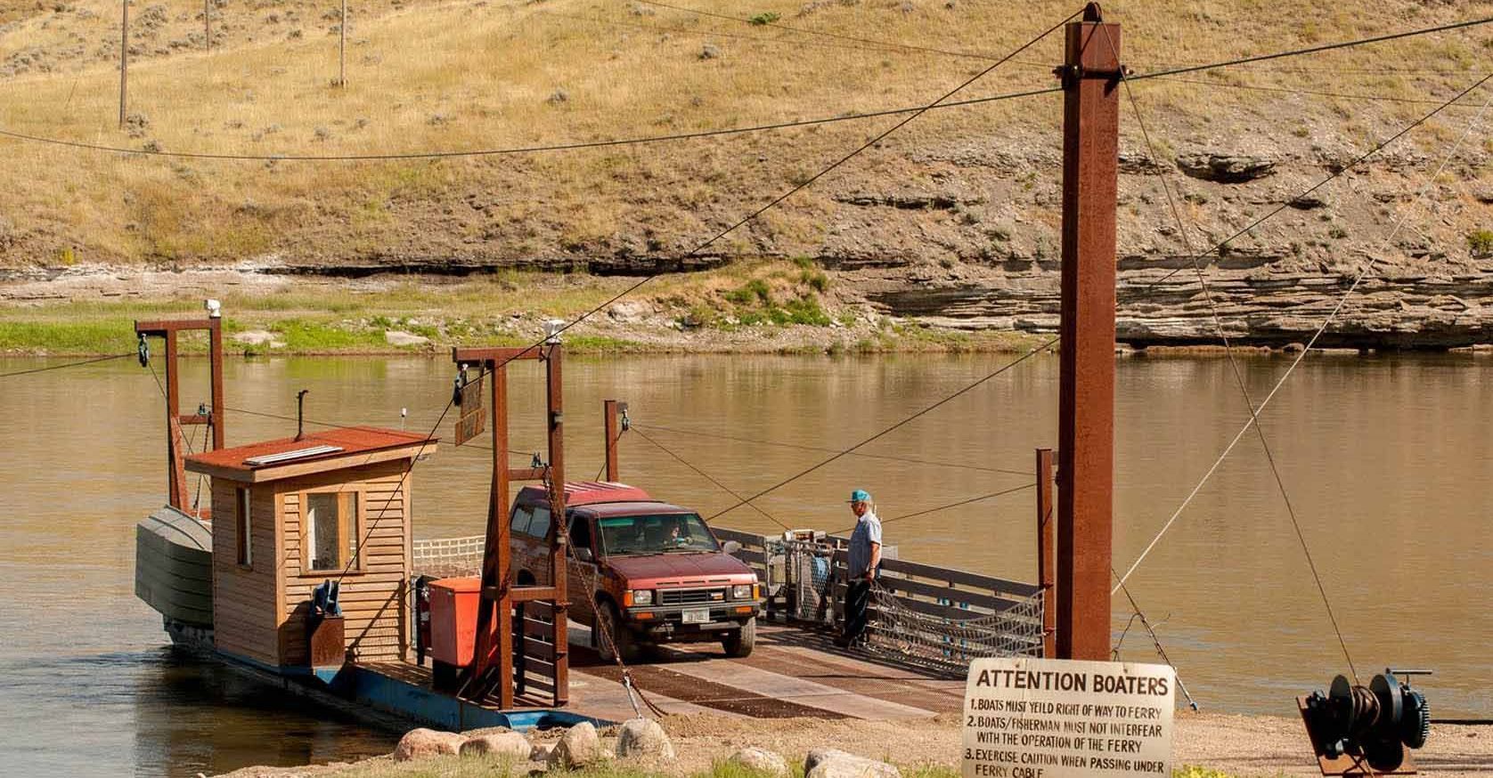 A red truck is being towed by a boat across a river.