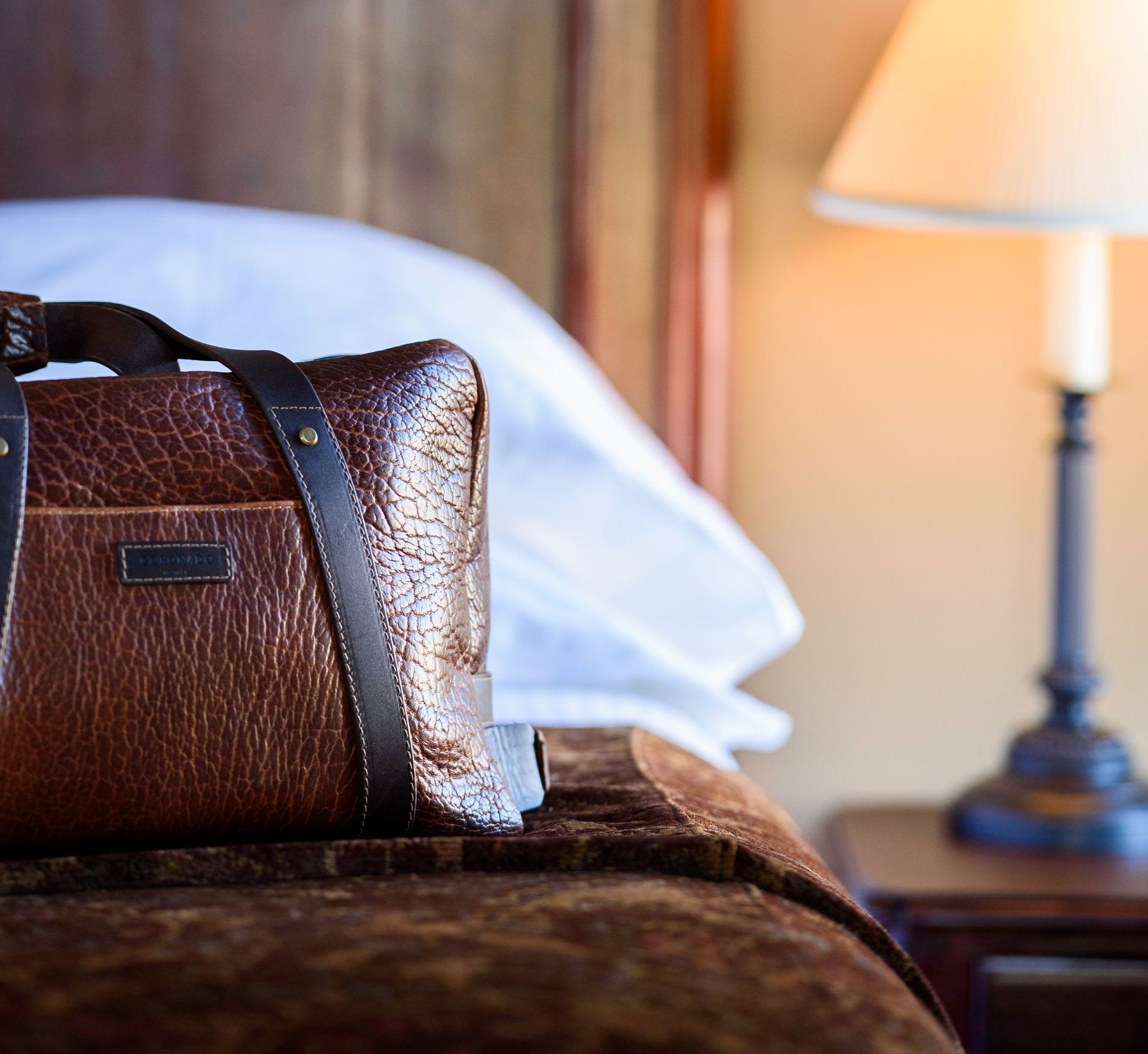A brown bag is sitting on a bed next to a lamp.