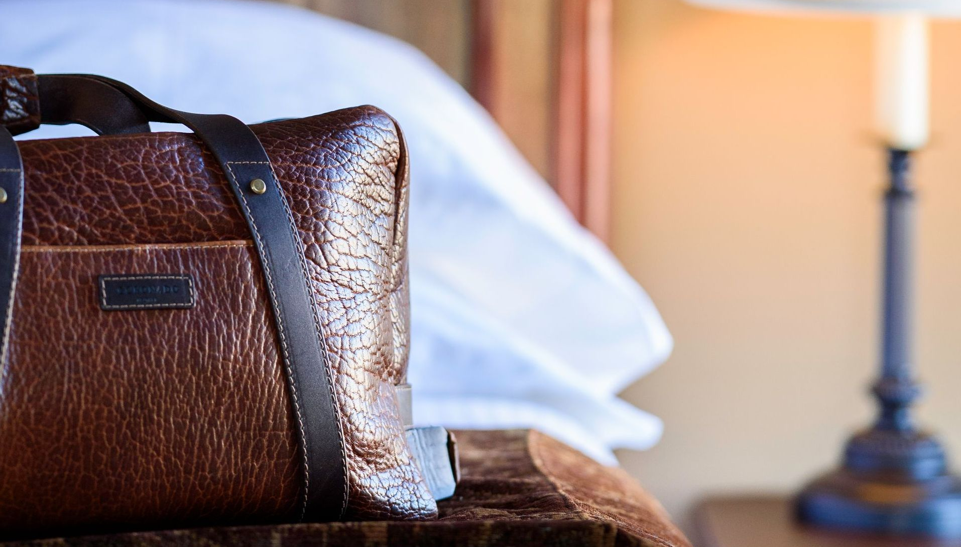 A brown bag is sitting on a bed next to a lamp.