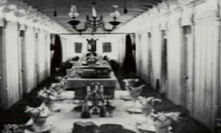 A long black and white photo of a dining room with a long table and chairs.
