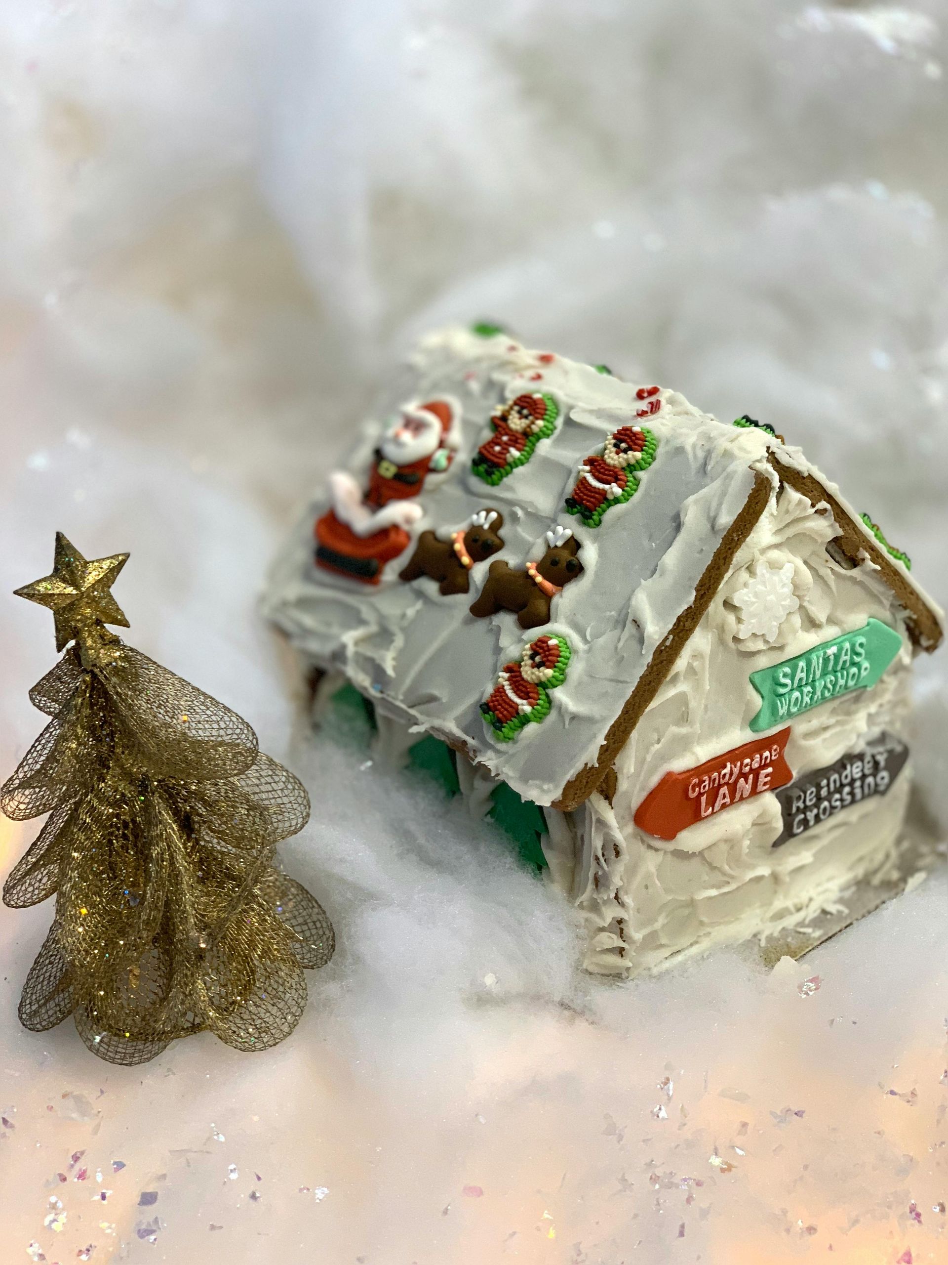 A gingerbread house decorated with santa claus and reindeer is sitting next to a christmas tree.