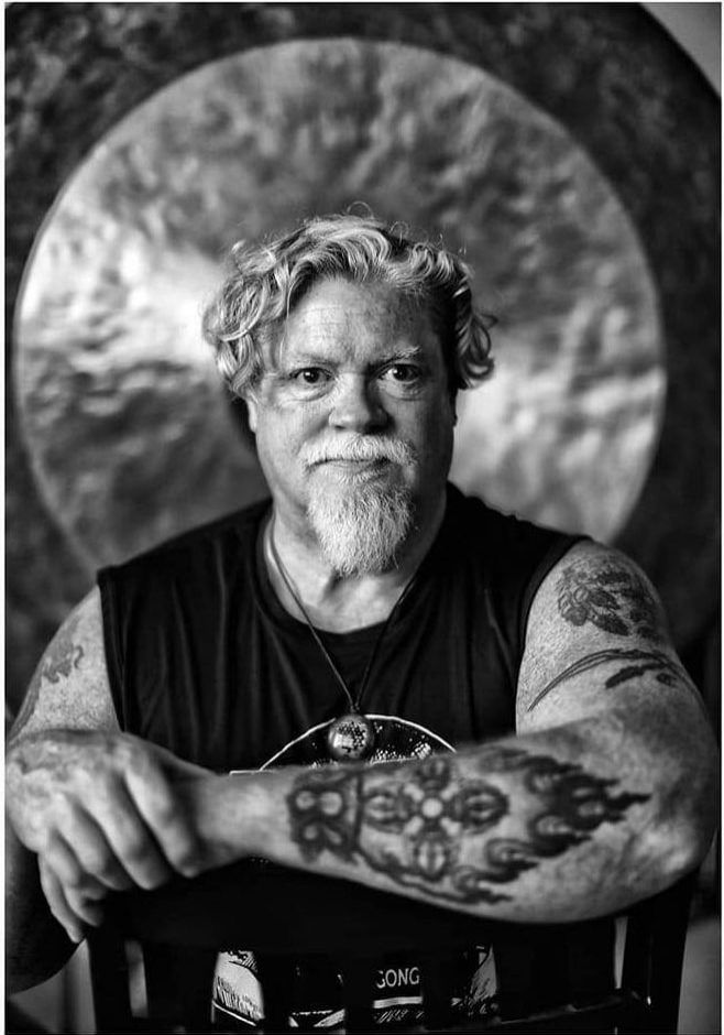 Man with tattoos and beard, sits before large gong.