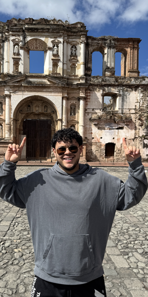 Man pointing up with both hands, in front of an old building. He wears sunglasses and a grey hoodie. Stone pathway.