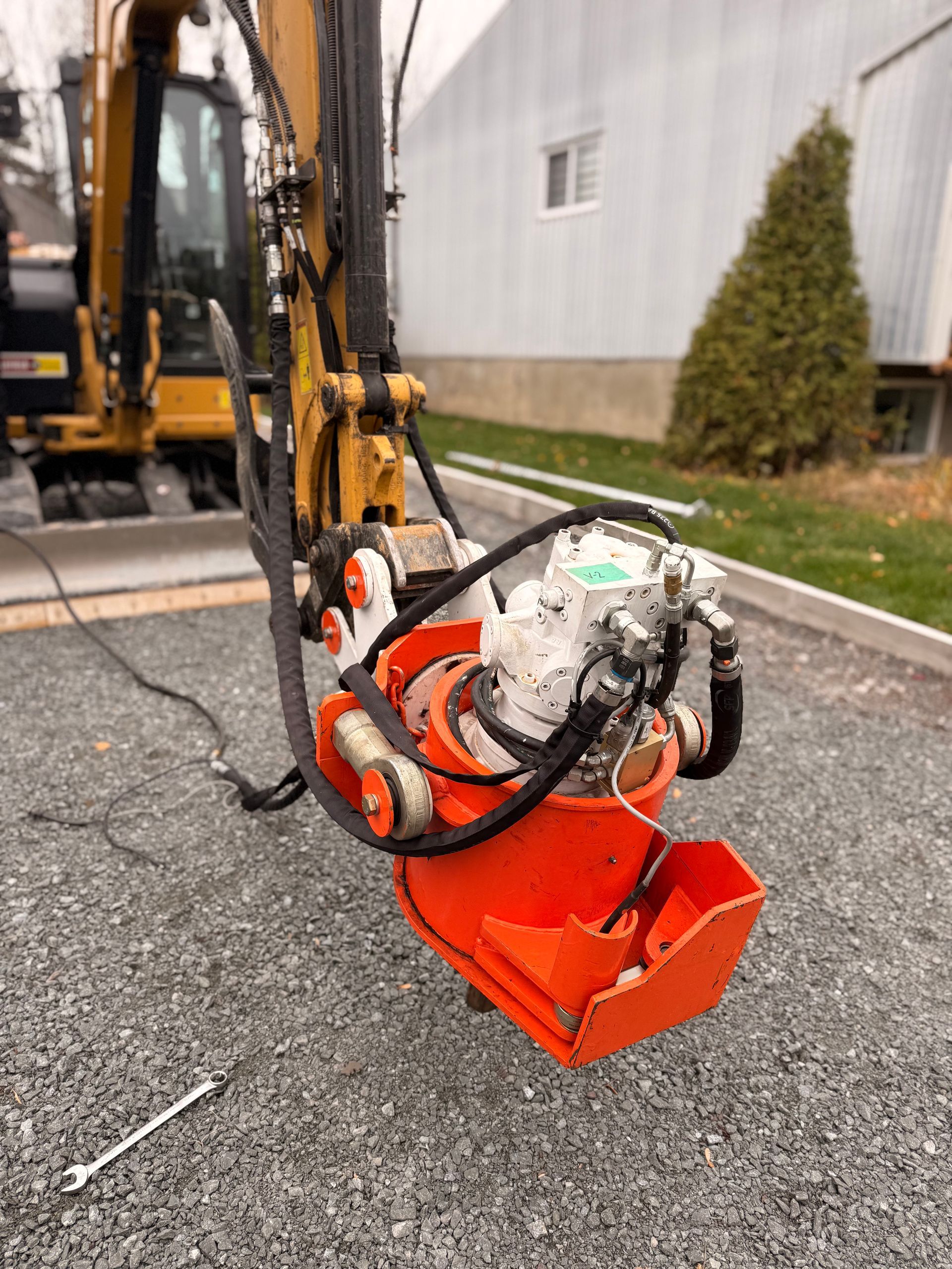 Accessoire d'excavatrice orange sur du gravier gris. Bras d'excavatrice jaune en arrière-plan, bâtiment blanc.