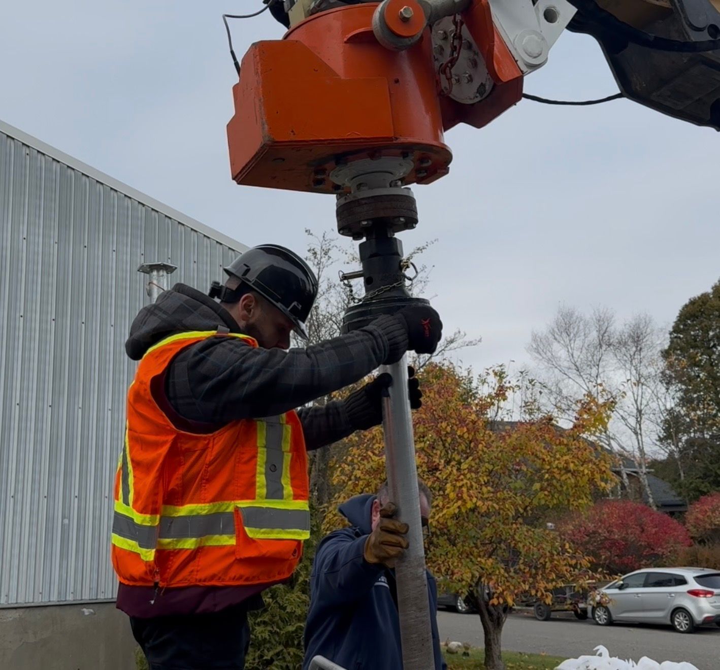 Un ouvrier en gilet orange fixe un poteau à une foreuse ; ciel couvert.
