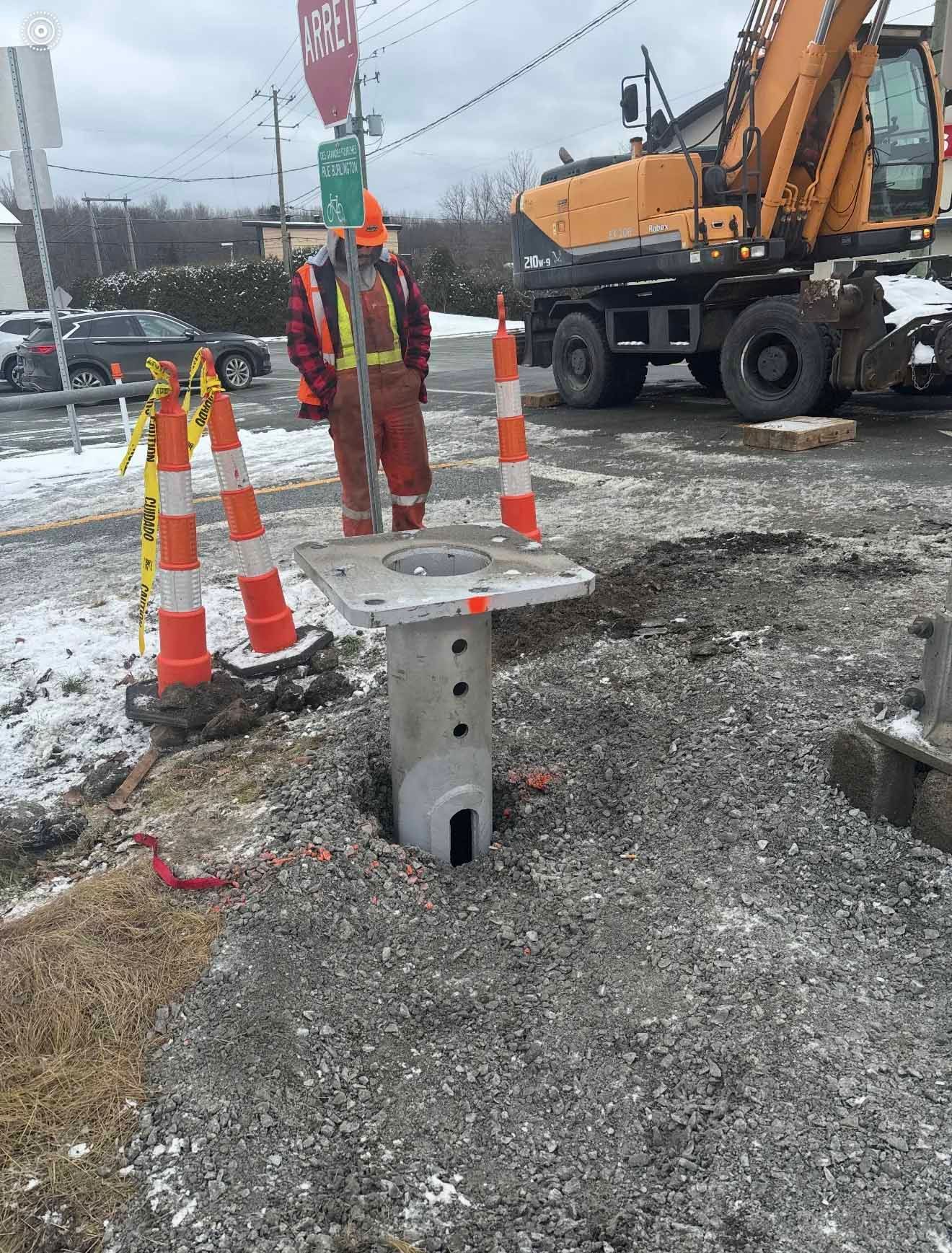 Ouvrier du bâtiment près d'un poteau électrique sur lequel on travaille à proximité d'une excavatrice. Cônes de sécurité orange. Zone de gravier enneigée.