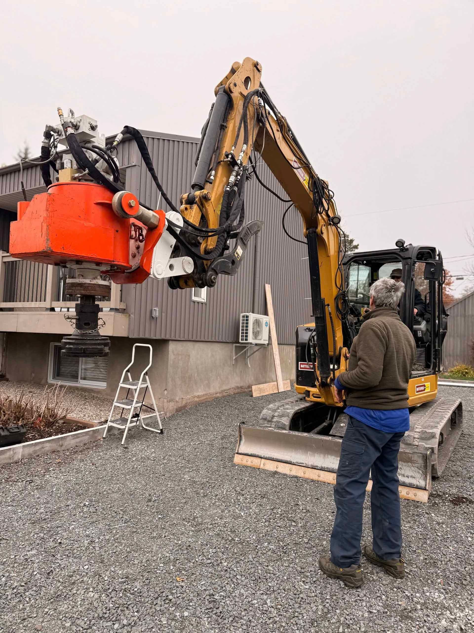 Un homme manœuvre une excavatrice équipée d'une pince près d'un bâtiment. Sol caillouteux. Ciel couvert.