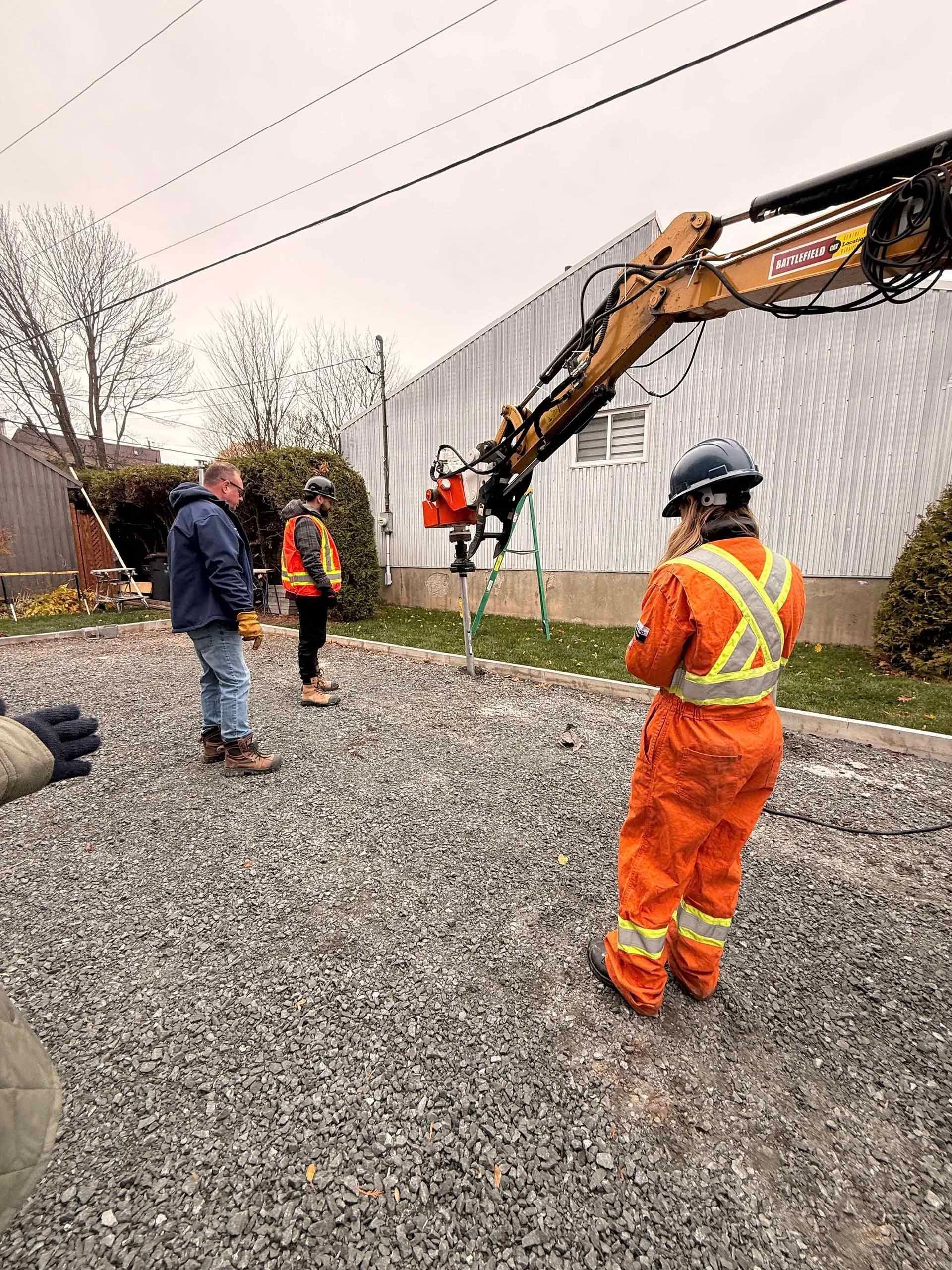 Des ouvriers équipés de protections manipulent des engins à proximité de lignes électriques. Surface gravillonnée. Ciel couvert.
