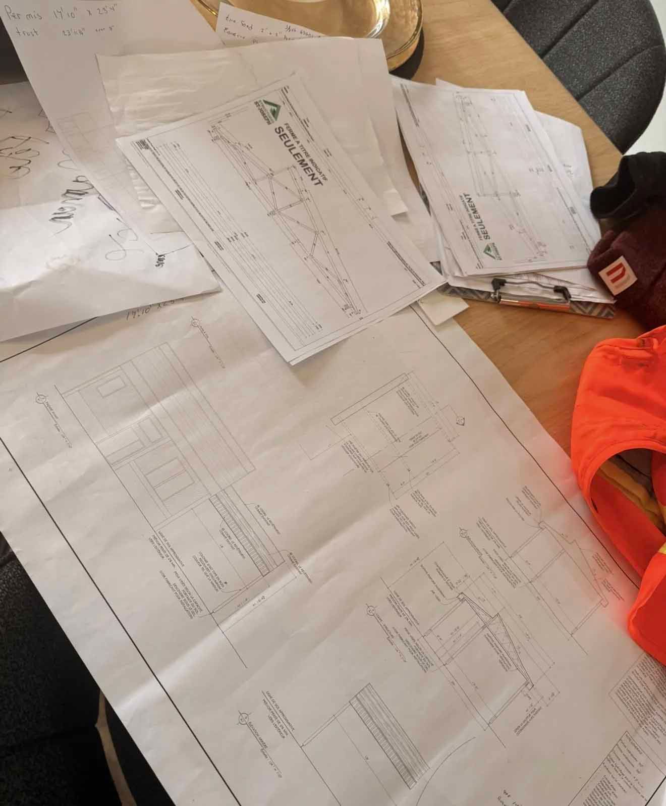 Des plans de construction étalés sur une table, des gants de travail et un gilet de sécurité à proximité.