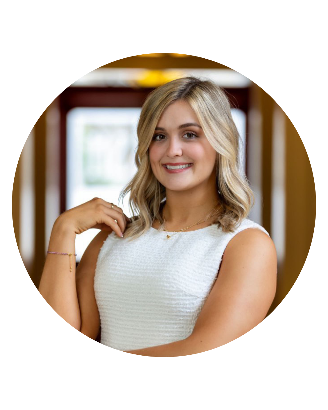 Woman with blonde wavy hair smiles, wearing a white sleeveless dress; indoors.