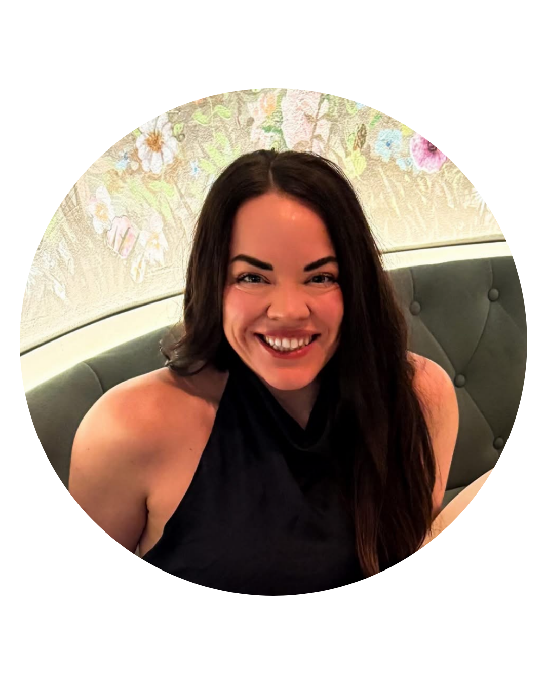 Woman with dark hair smiles, wearing a black halter top. Floral wallpaper backdrop.