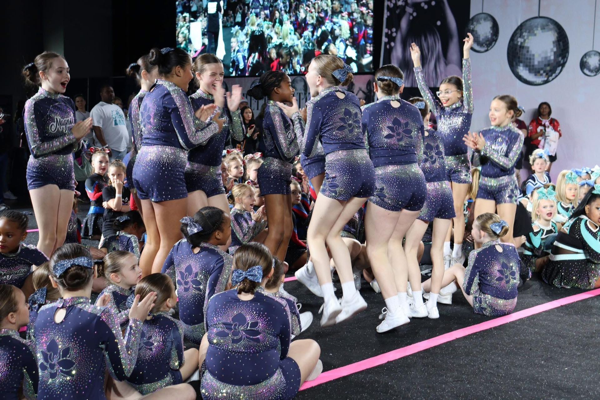 Cheerleaders in navy uniforms celebrate on stage, clapping and jumping. Sparkly accents and a crowd in the background.