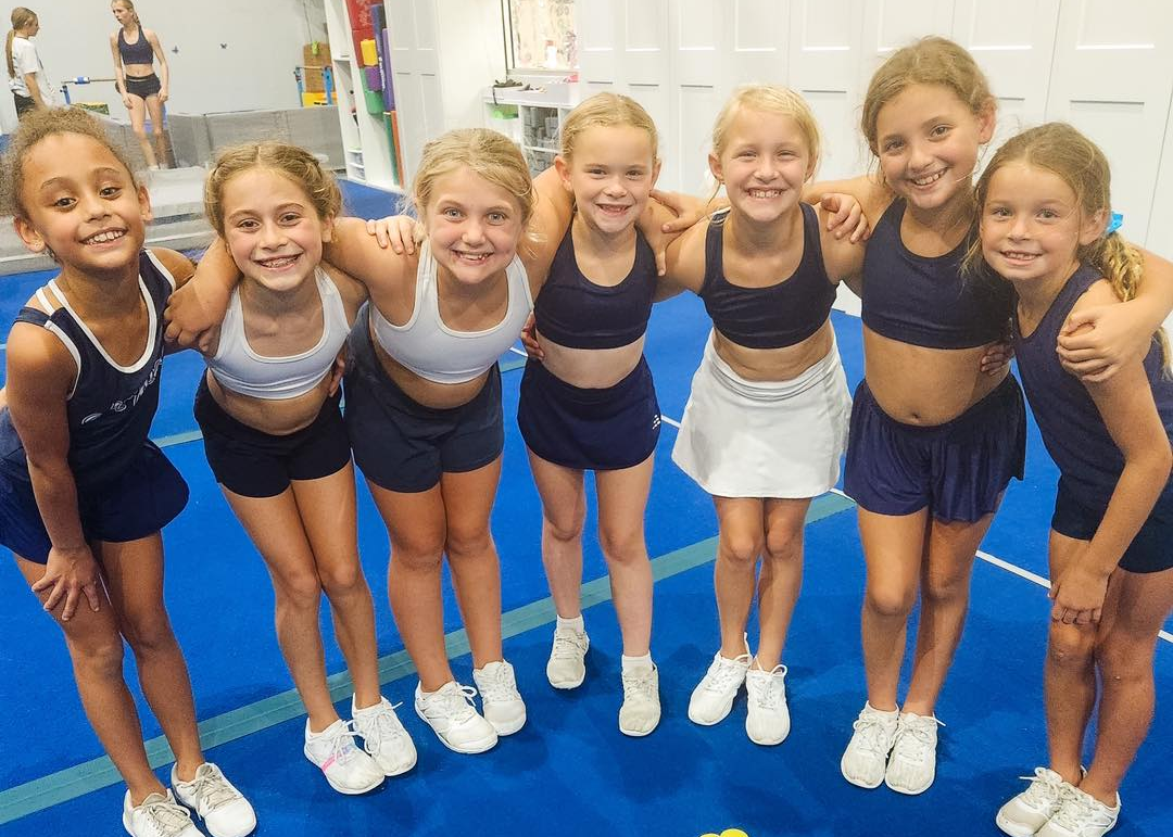Group of young cheerleaders in uniform, smiling and hugging, in a gym.