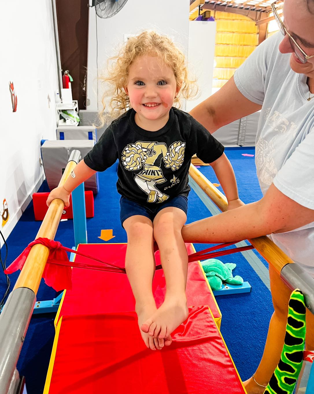 Young child smiling, on gym equipment, being assisted by an adult.