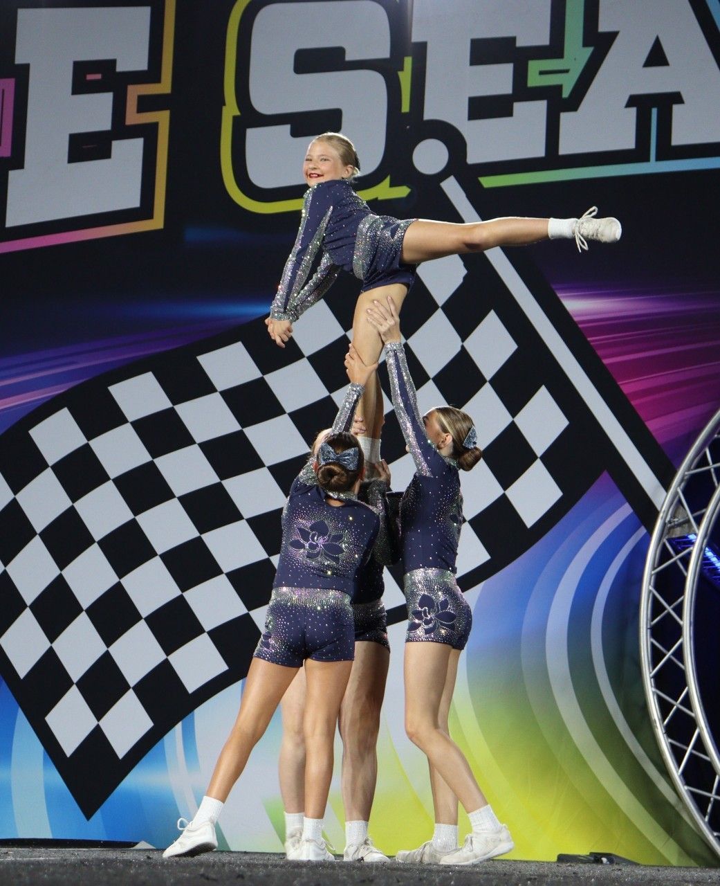 Cheerleaders in navy blue outfits perform a stunt, one person in the air with leg extended, set against a stage backdrop.