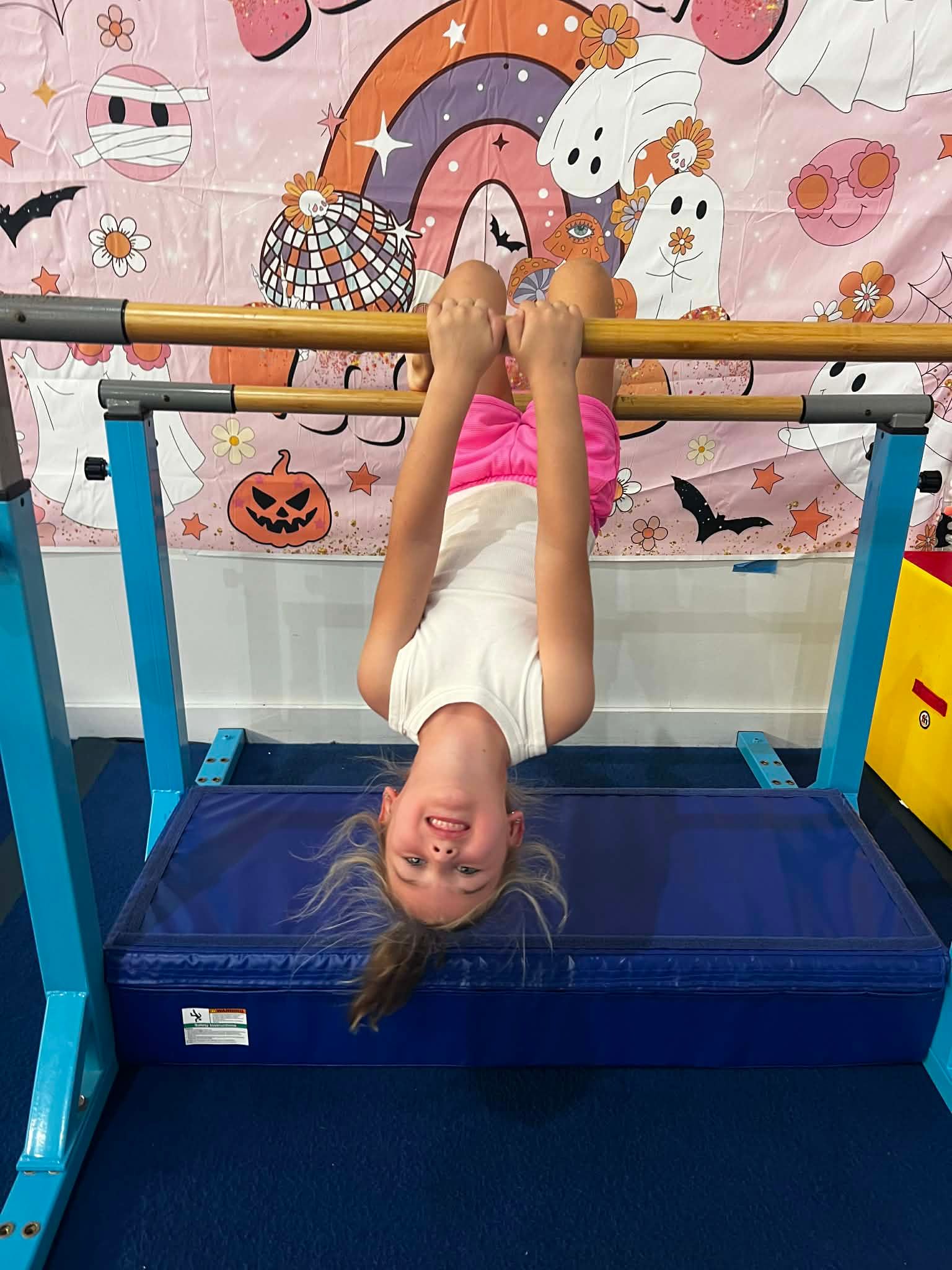 Girl smiling upside down, hanging on gymnastics bar above a blue mat; Halloween-themed backdrop.
