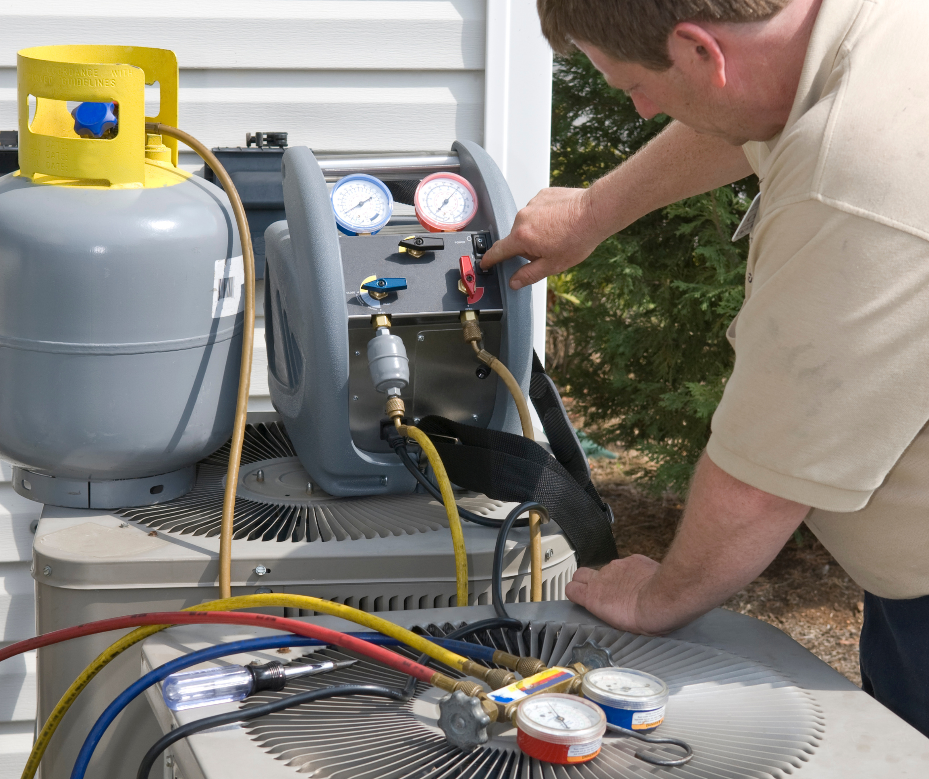 A technician working on an AC unit, connecting hoses to a gauge, next to a refrigerant tank.