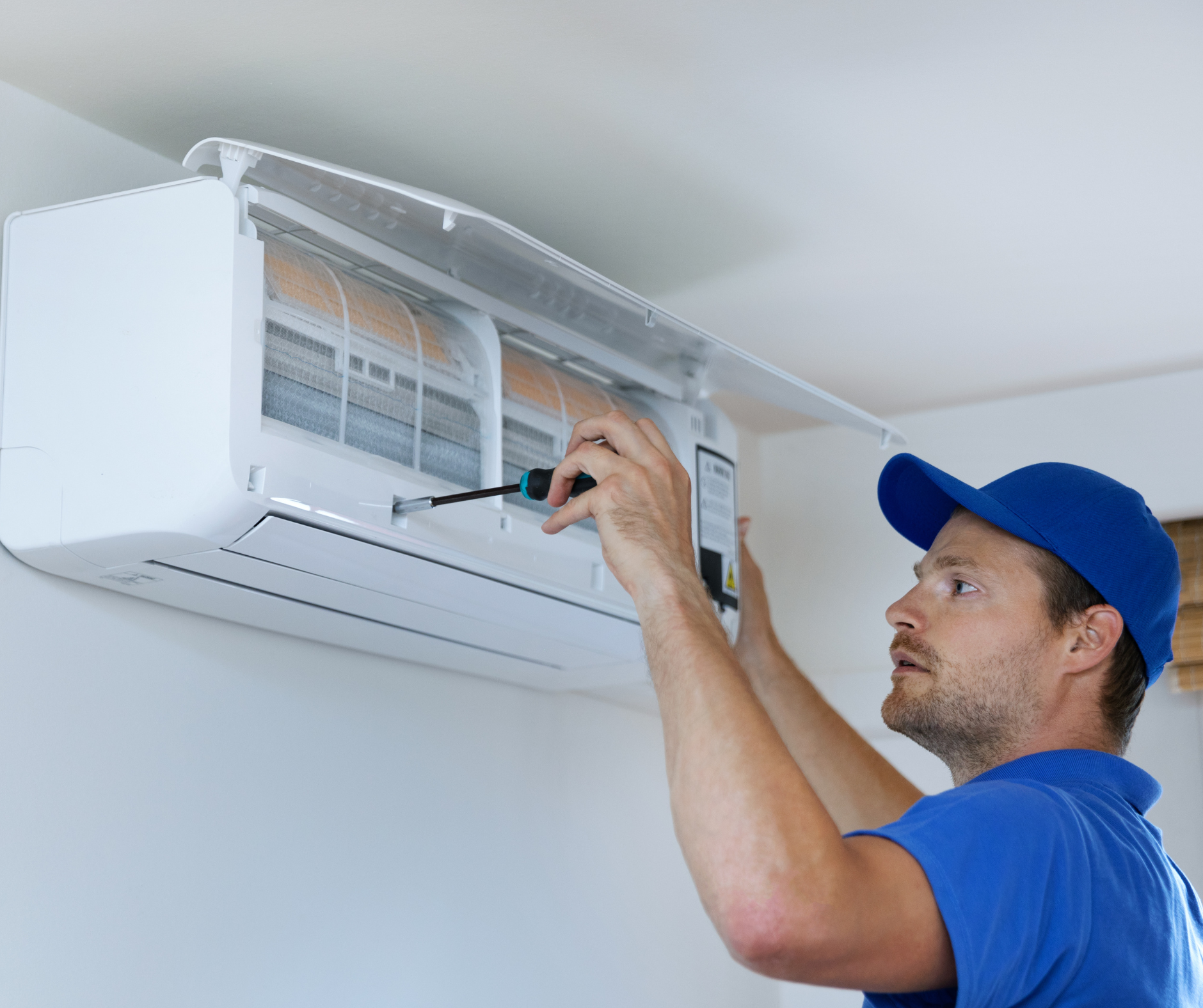 HVAC technician in blue uniform servicing a white wall-mounted air conditioner with a screwdriver.